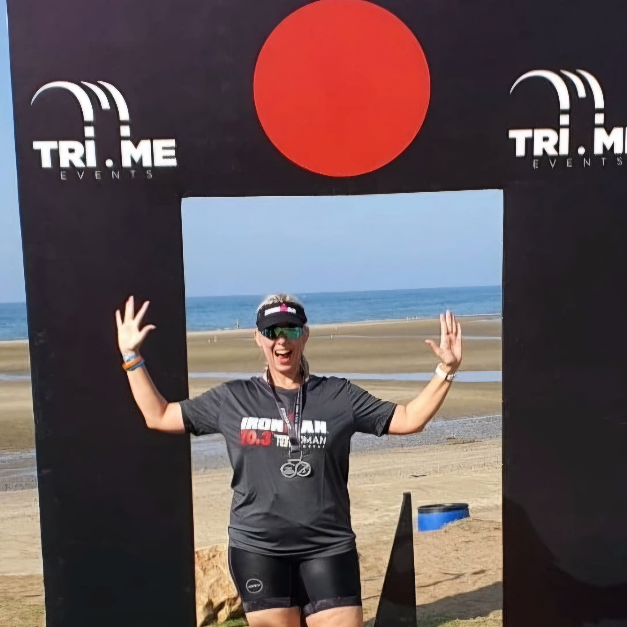 A woman celebrating at the finish line of a triathlon event on a beach, standing under a black arch with a red circular logo and the text 'TRI ME EVENTS,' with the ocean in the background.