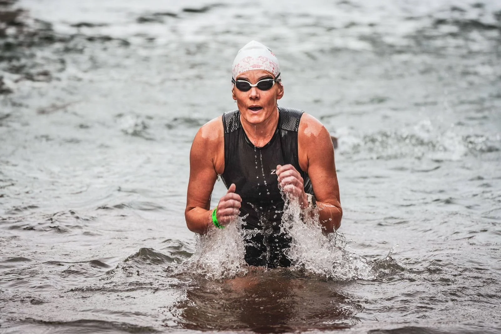 An older woman wearing a swim cap, goggles, and a black athletic top, running out of water in a triathlon or open-water swim event.