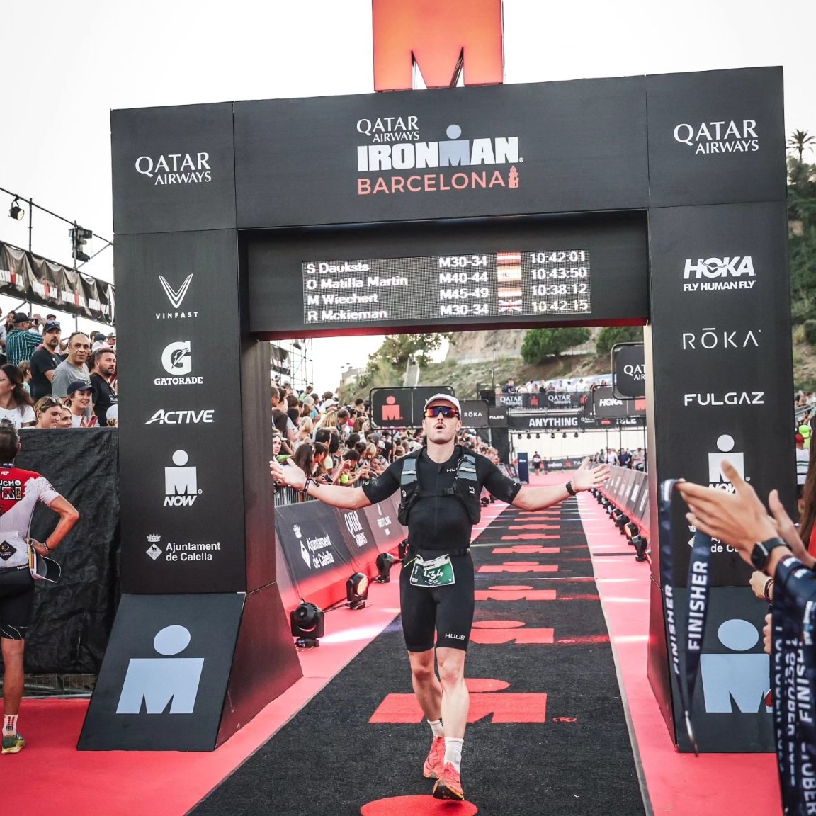 Male triathlon finisher crossing the finish line at Ironman Barcelona with arms outstretched and celebration expression, surrounded by crowd and race banners.