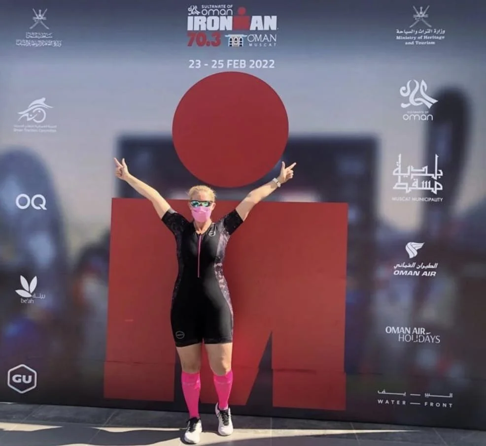 A woman in a black and pink sports outfit, pink knee-high socks, and sunglasses, standing in front of a large promotional banner for Ironman 70.3 Oman, dated February 23-25, 2022, with her arms raised in a celebratory pose.