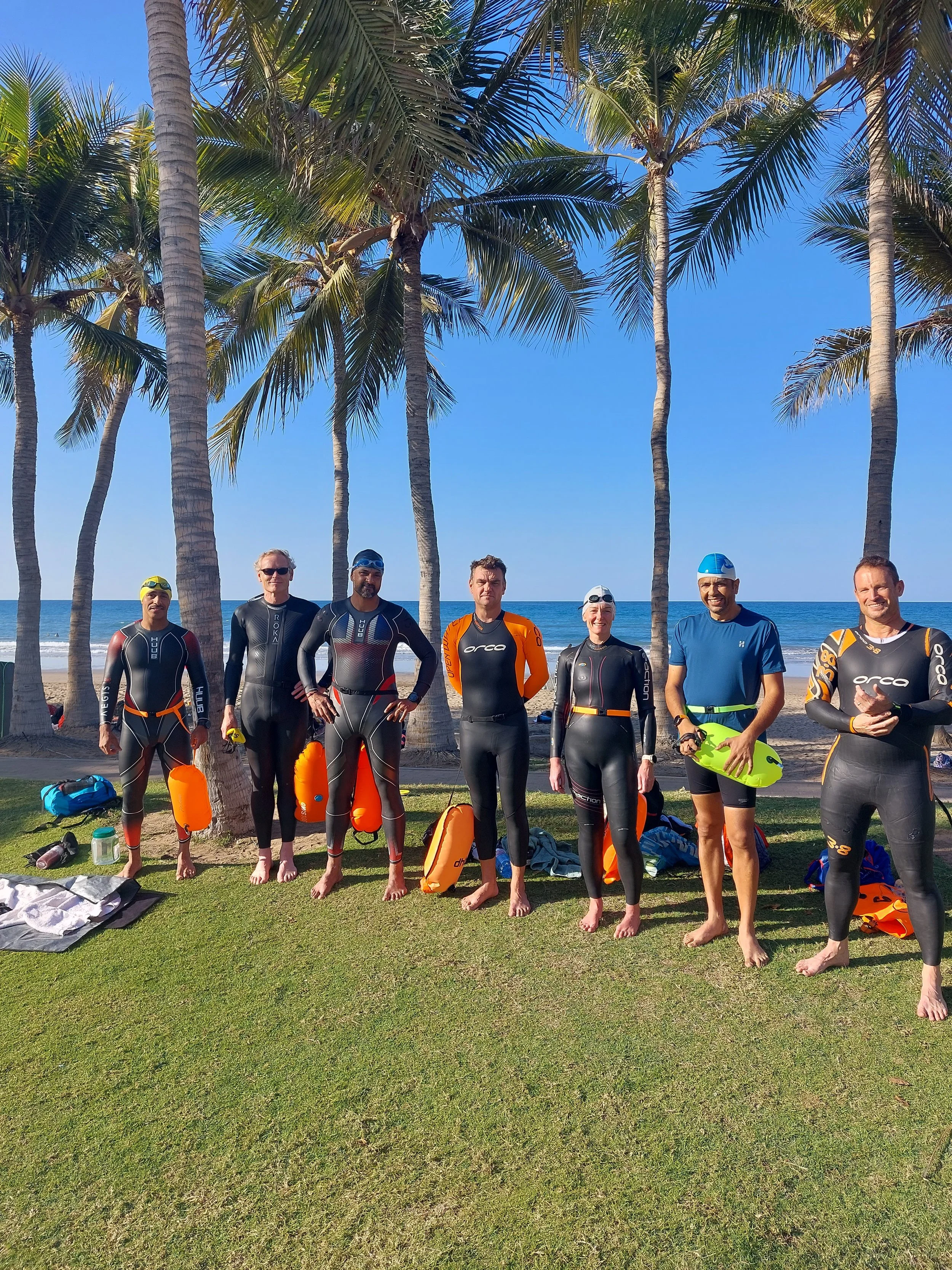 Group of eight people in wetsuits standing on grass near palm trees at the beach, with the ocean and blue sky in the background.