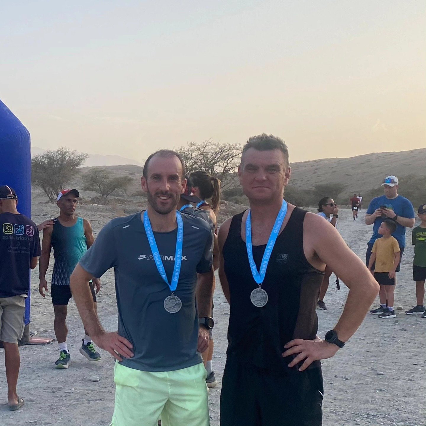 Two men wearing medals and athletic clothing posing at the finish line of a race in a desert landscape, with other runners and spectators in the background.