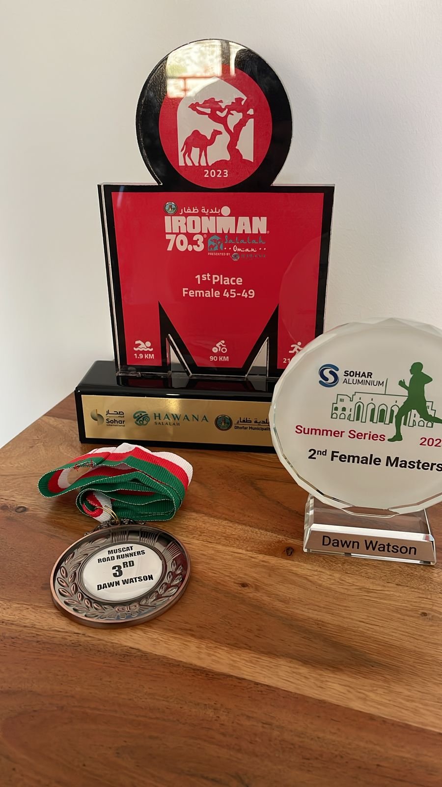 Trophies and medals from a running race. The main trophy says '1st Place Female 45-49' for the Ironman 70.3 event, with race distances of 1.9 km swim, 90 km bike, and 21 km run. A second trophy states '2nd Female Master' from the Sohar Aluminium Summer Series 2023. A bronze medal with a green, red, and white ribbon reads '3rd Muscat Road Runners Dawn Watson.'
