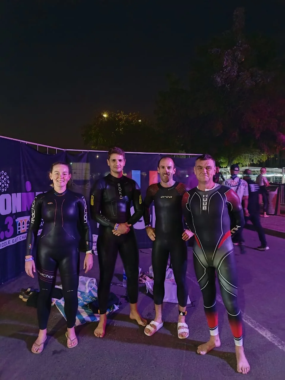 Four athletes in wetsuits standing together outdoors at night, smiling, after a swimming event.
