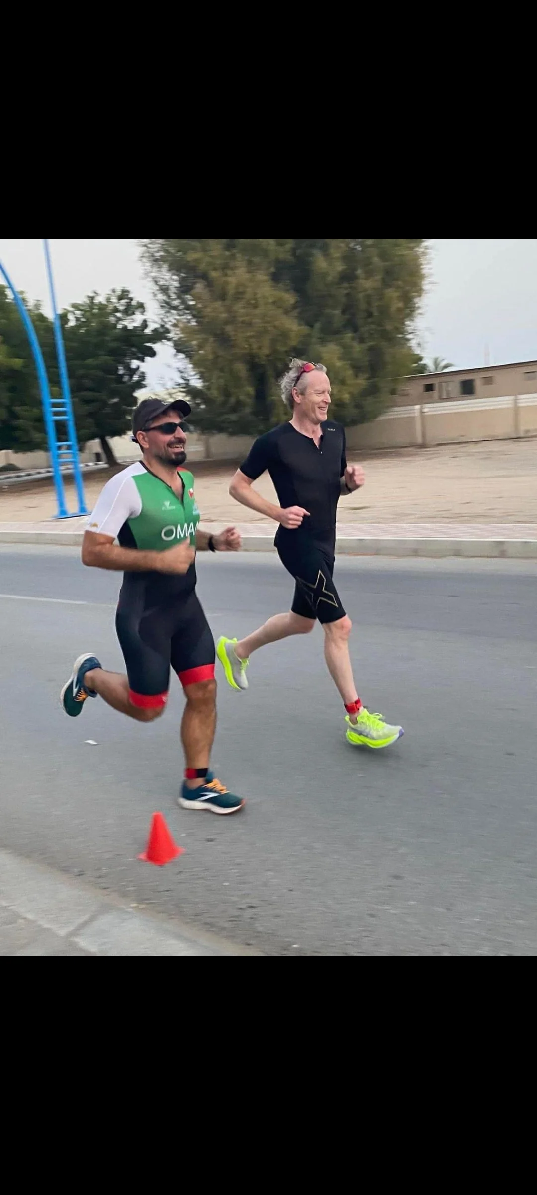 Two men running on a street during a race, with one wearing black and yellow athletic gear and the other in a black, green, red, and white outfit, smiling and enjoying the run, with orange traffic cones marking the course.