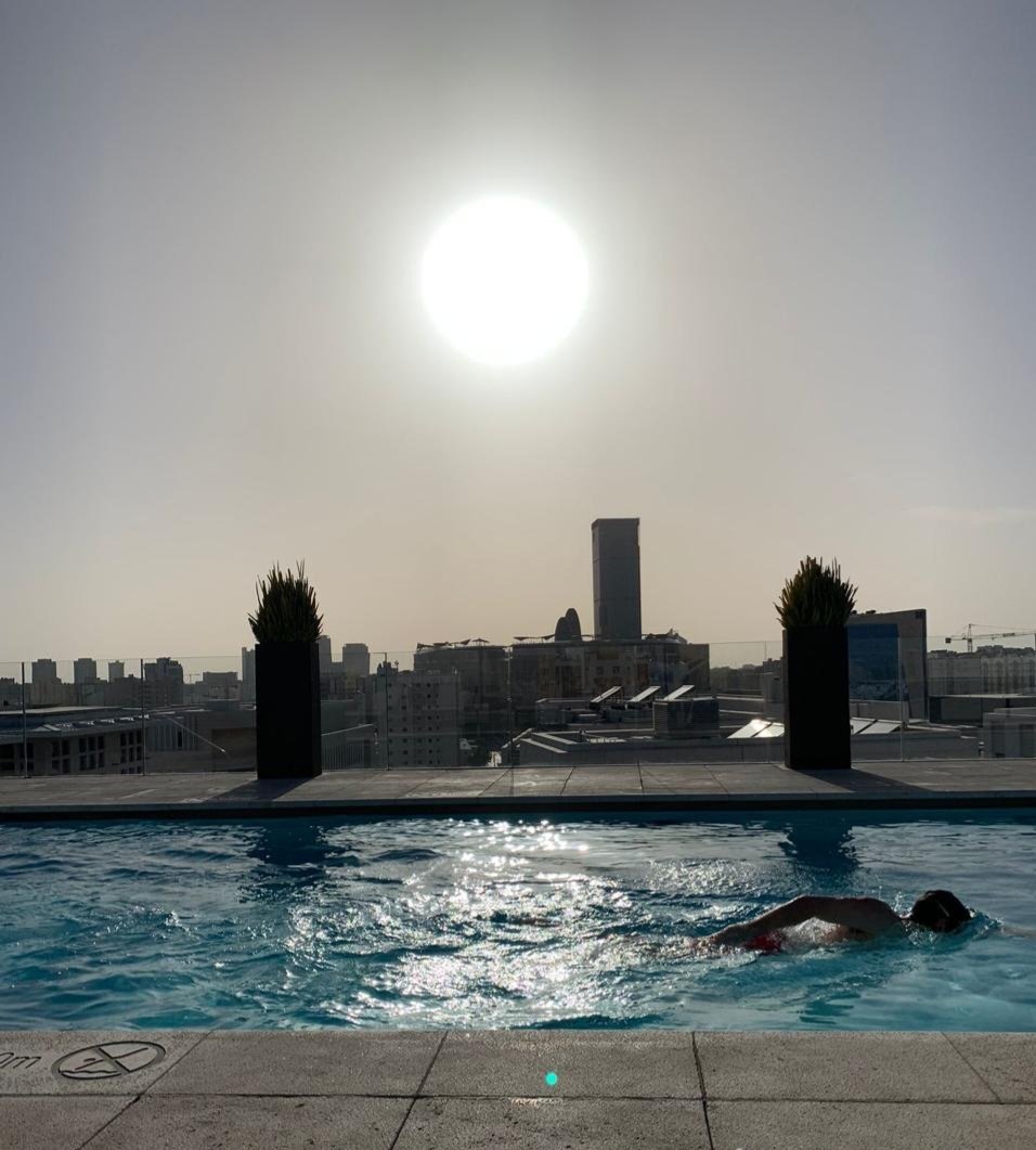 A person swimming in a rooftop pool with city buildings and a bright sun in the background.