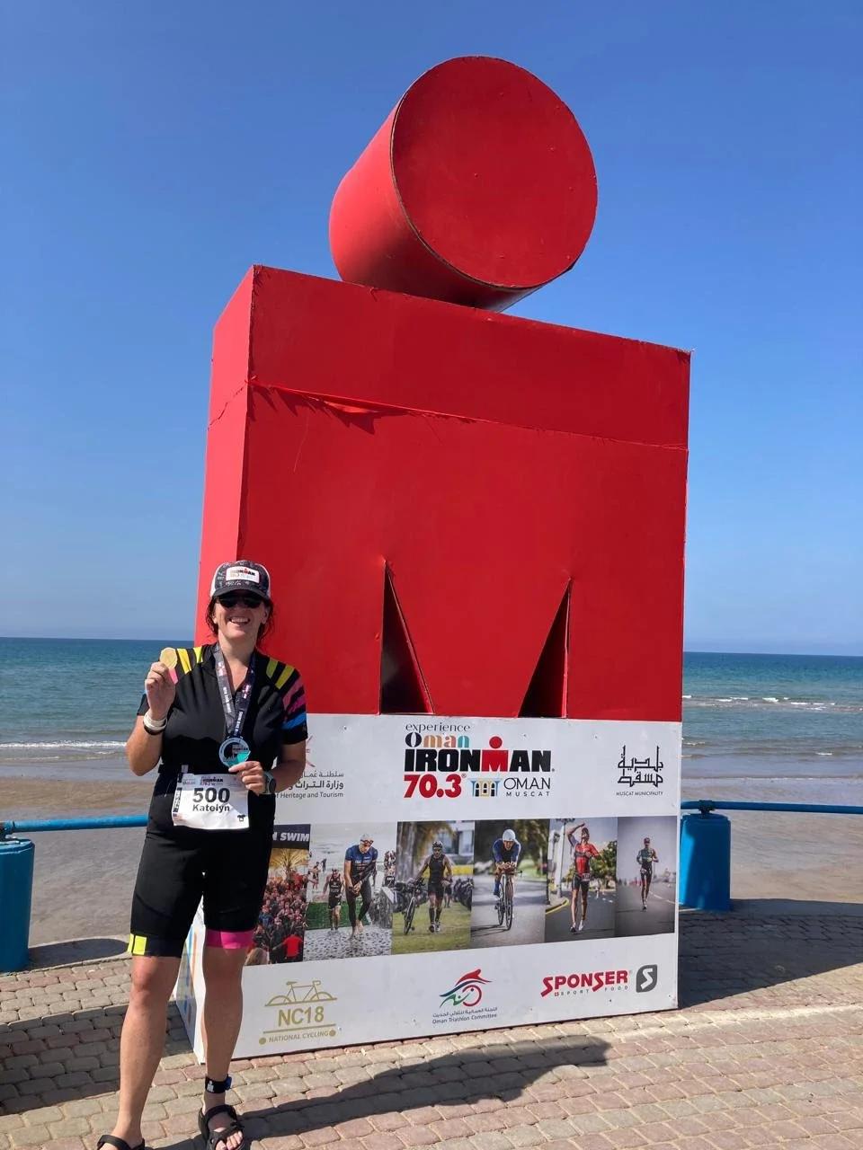 A woman in black athletic clothing holding a medal and smiling at a beachside background with a large red Ironman sign and sponsorship banners.
