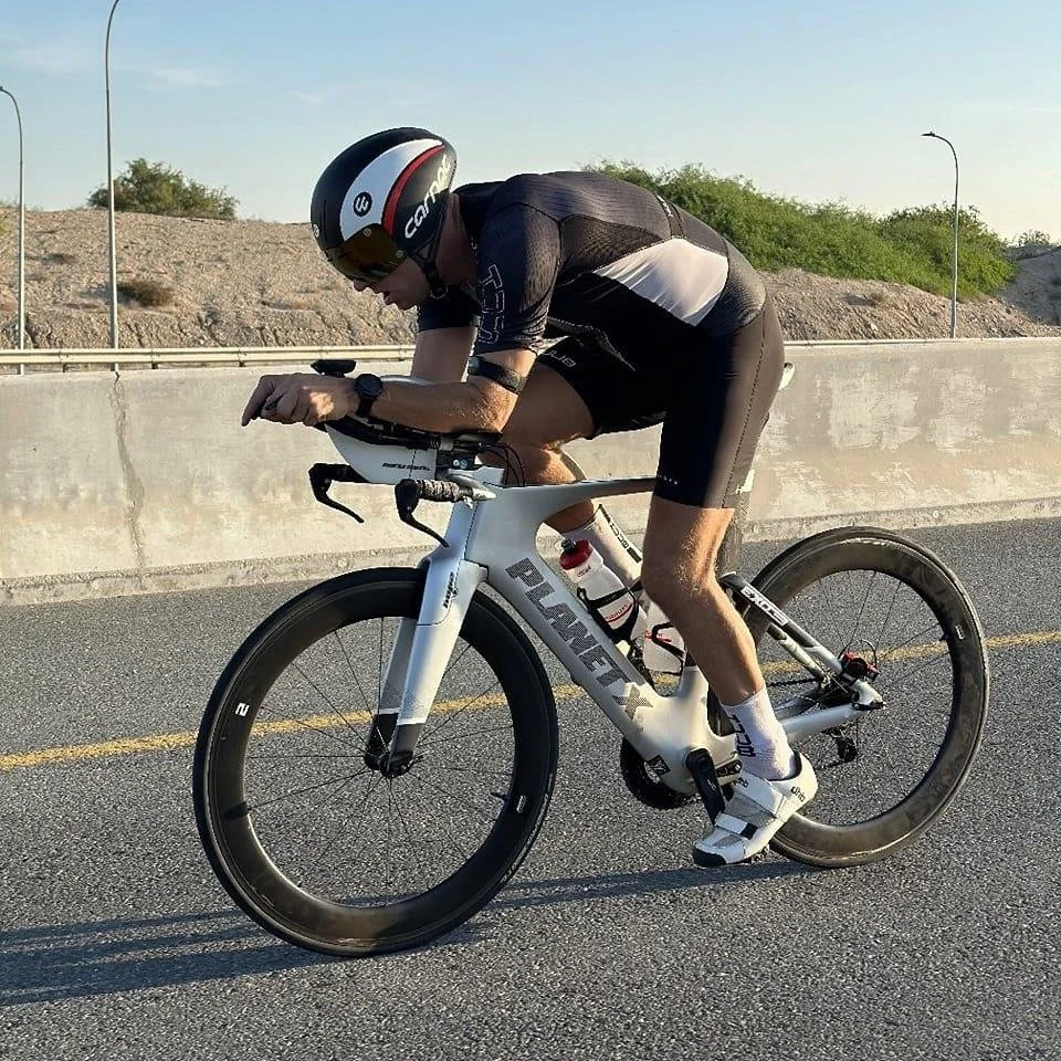 A cyclist wearing a black helmet and black riding suit bends forward on a white road bike, riding on a paved road with a concrete barrier and desert landscape with sparse vegetation in the background.