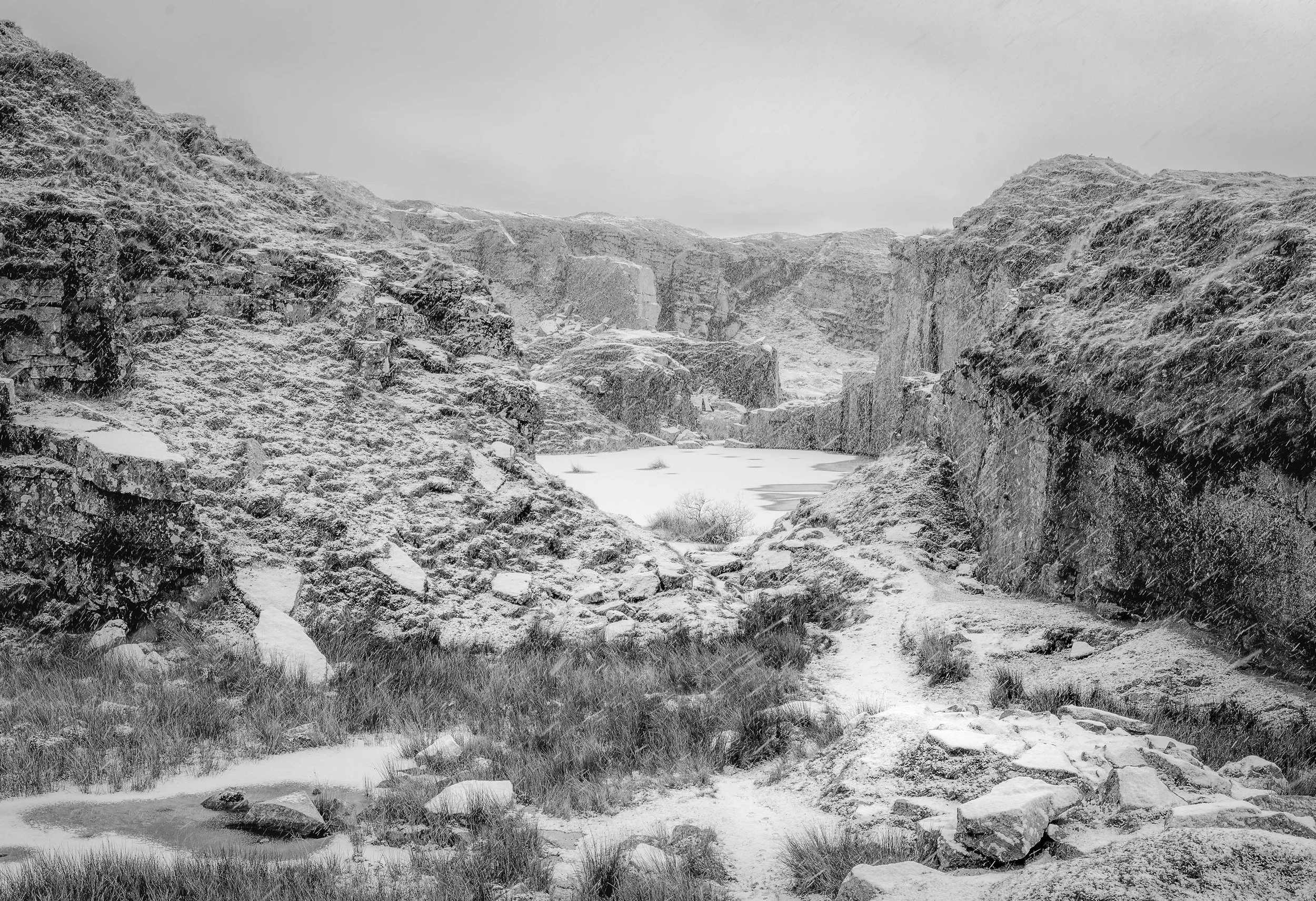 Snow-covered rocky canyon with a frozen pond in the middle and rugged hills on both sides.