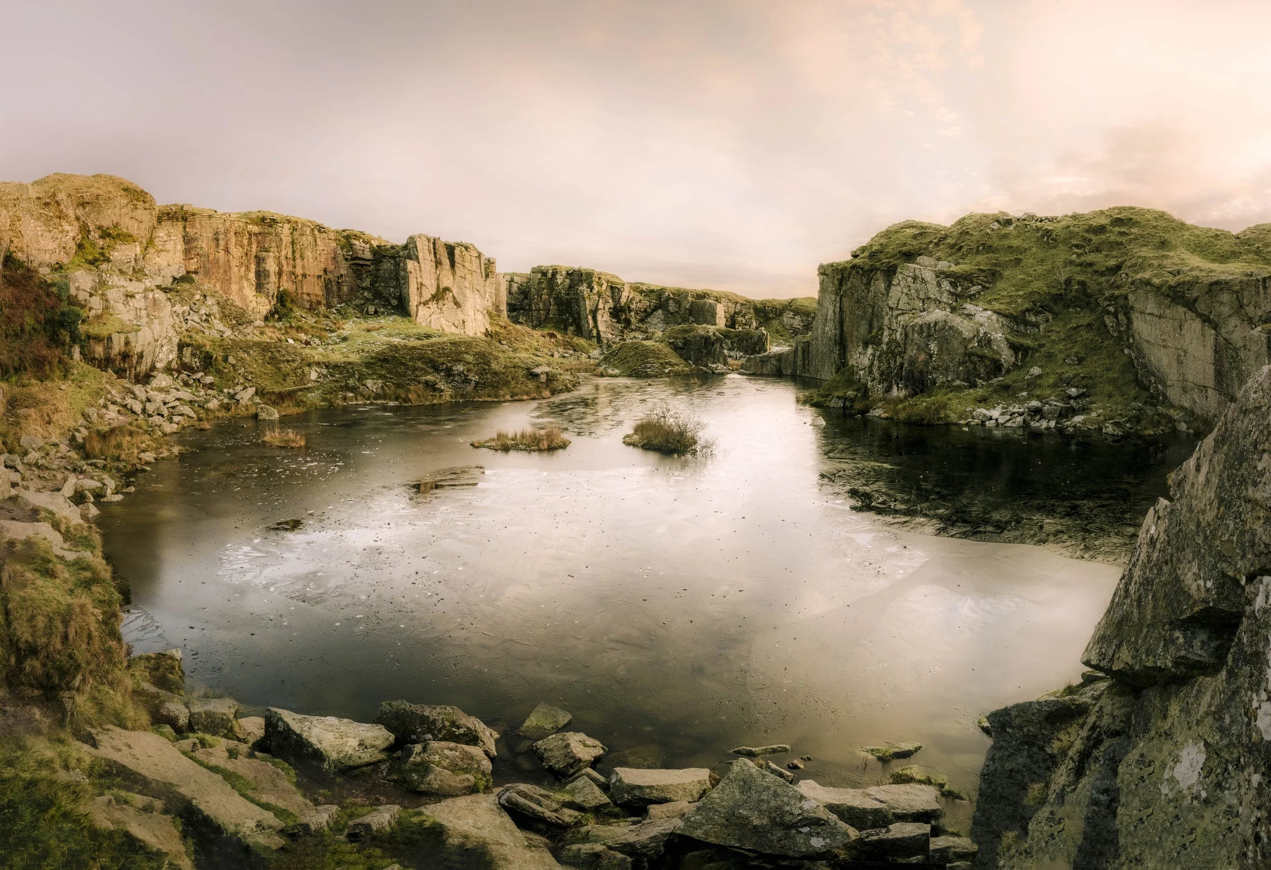 A river flows between rocky cliffs with sparse grassy patches, under a cloudy sky during sunset or sunrise.