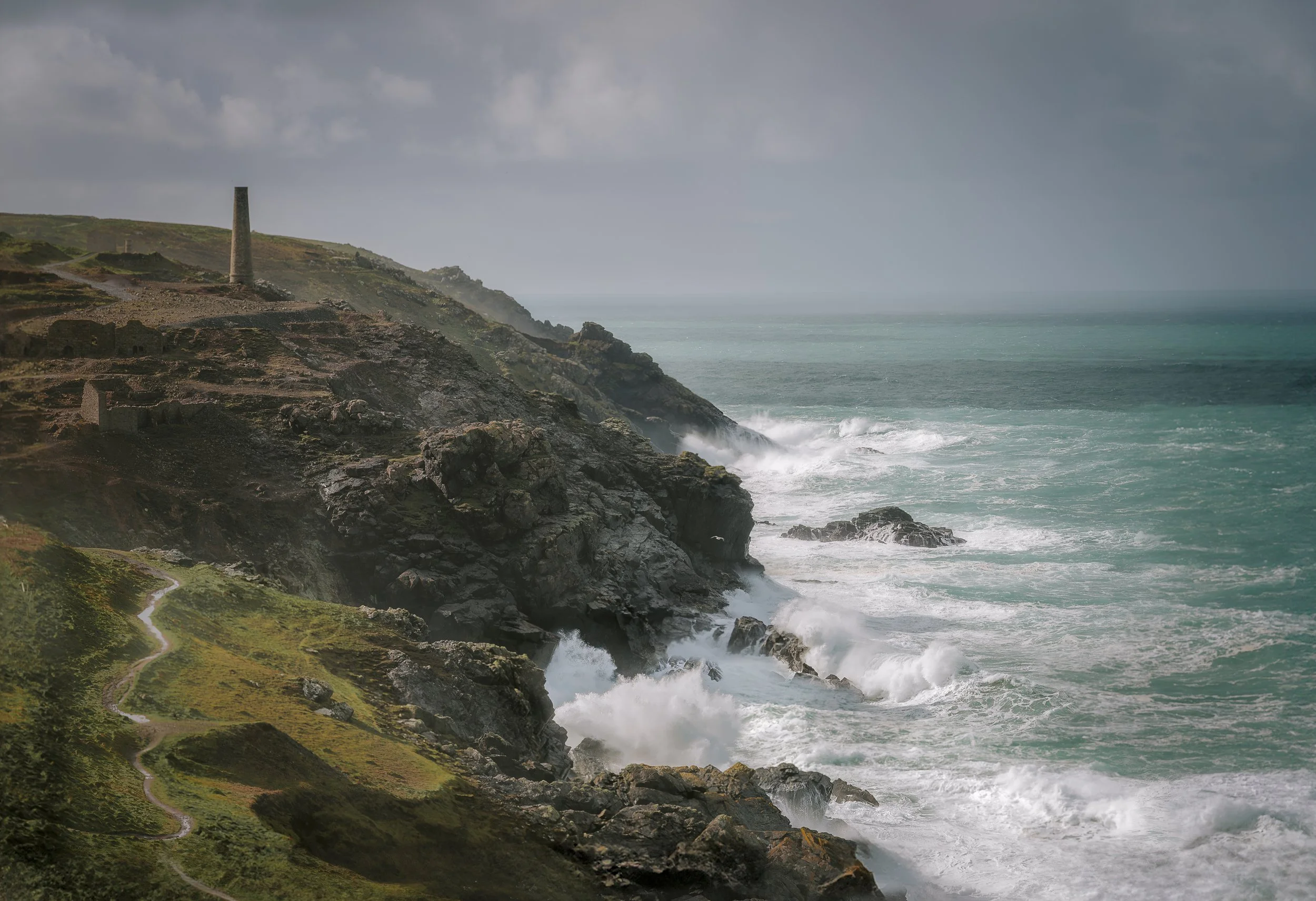 Coastal landscape with rocky cliffs, a lighthouse, a winding path, and ocean waves crashing against rocks under a cloudy sky.