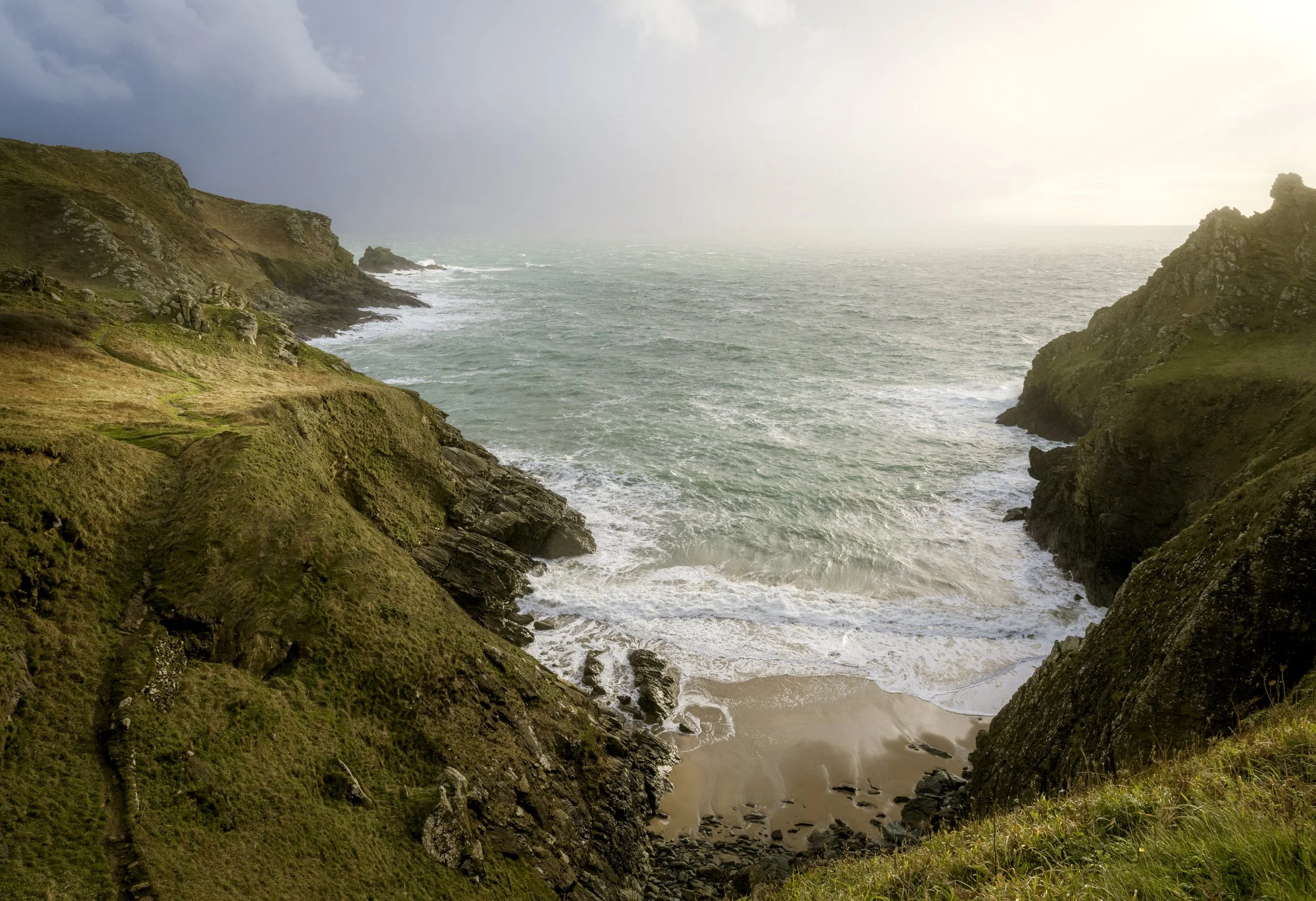 A coastal view of cliffs covered in green grass overlooking a sandy beach and ocean waves, under a cloudy sky with sunlight breaking through.