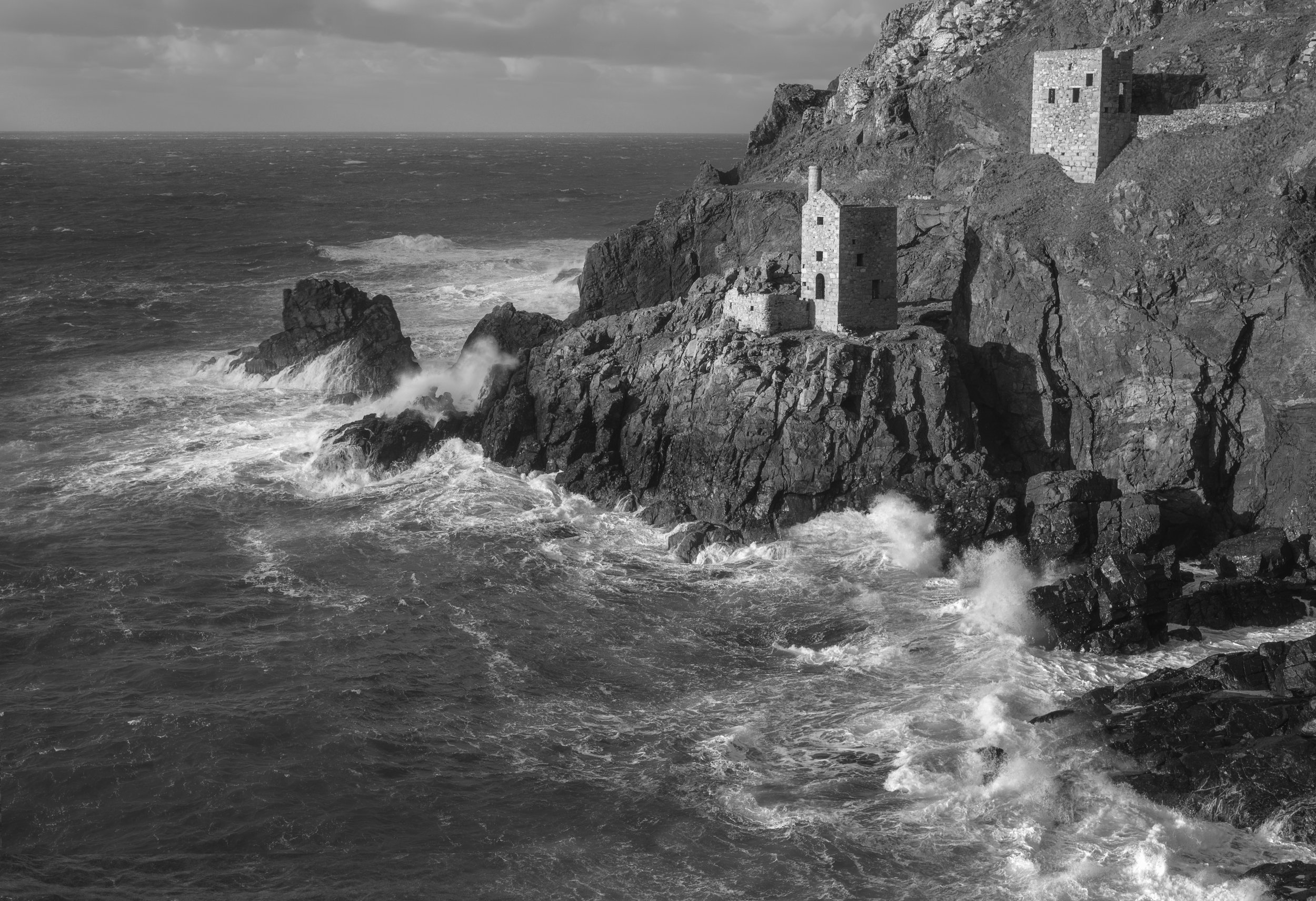 Black and white photo of a rocky coastline with two stone towers built on the cliff, waves crashing against the rocks below.