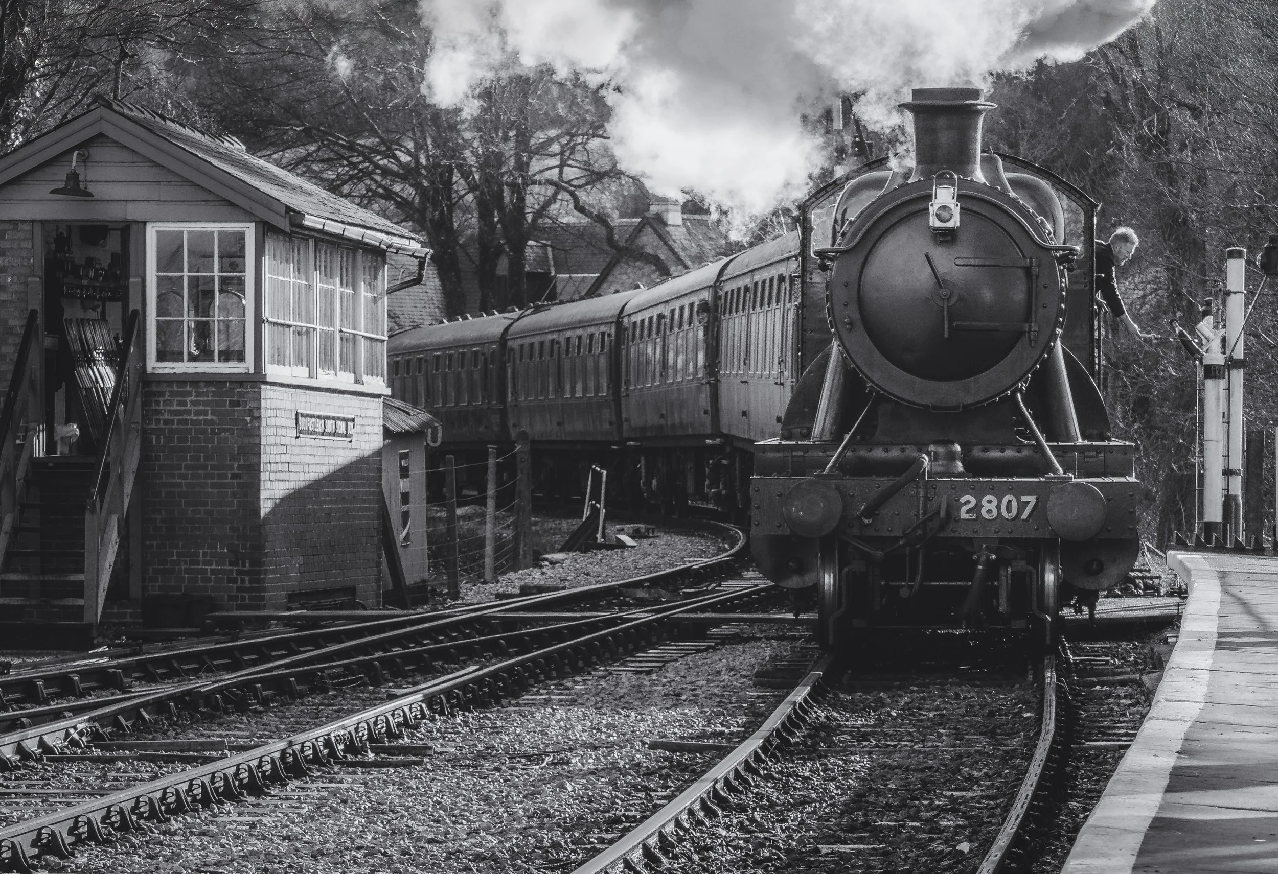 Black and white photo of a vintage steam locomotive train approaching a small train station with a platform and a signal arm, surrounded by trees in the background.
