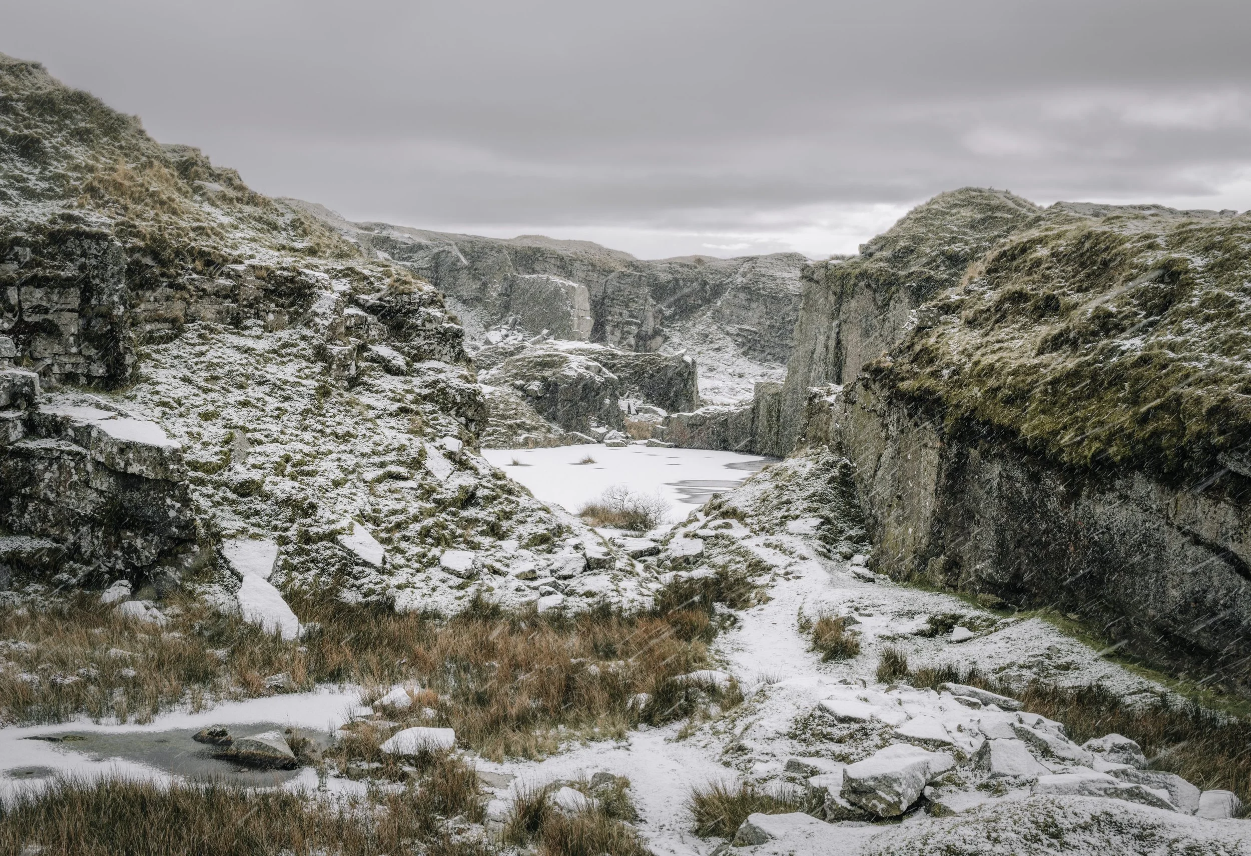 Snow-covered rocky landscape with a frozen body of water and mossy cliffs under an overcast sky.