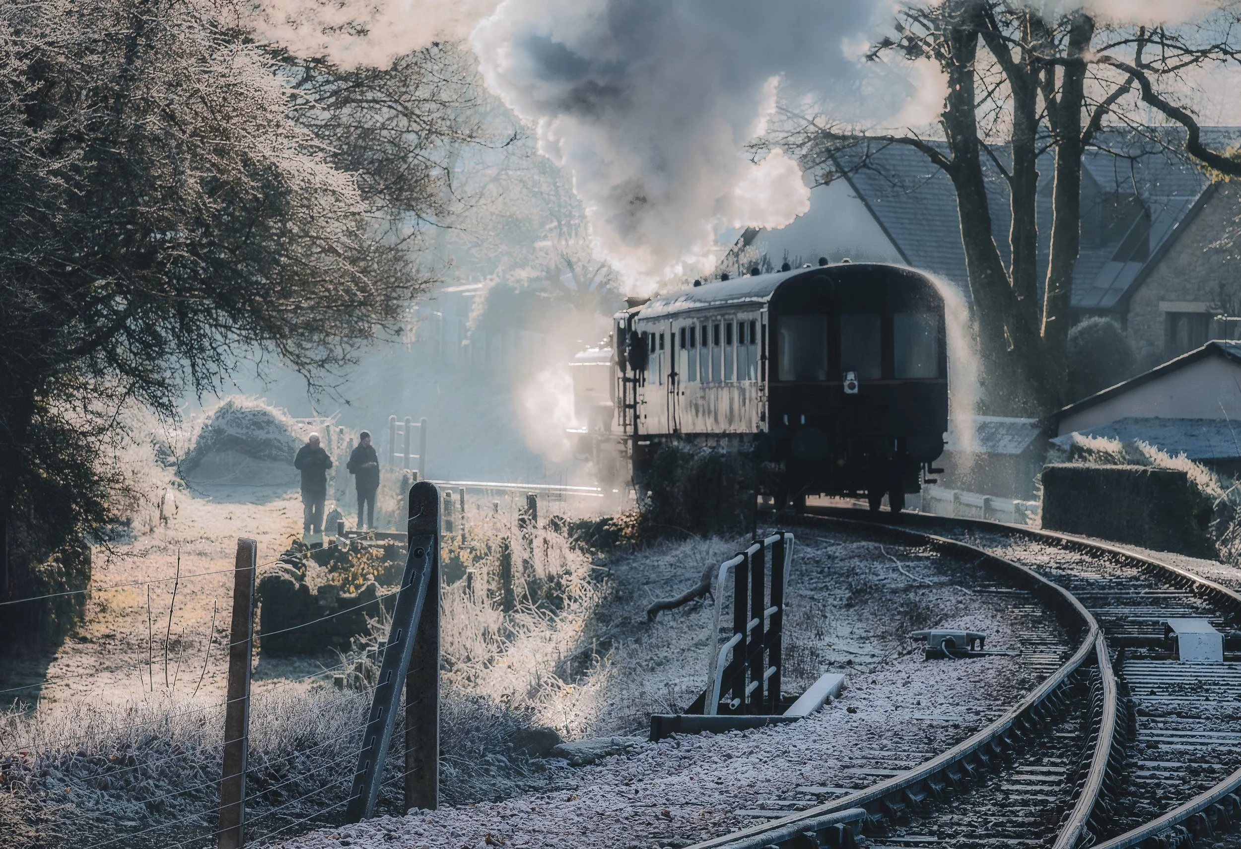 Steam locomotive train on snowy track passing through a winter landscape with trees and houses, with two people walking nearby on the left side.