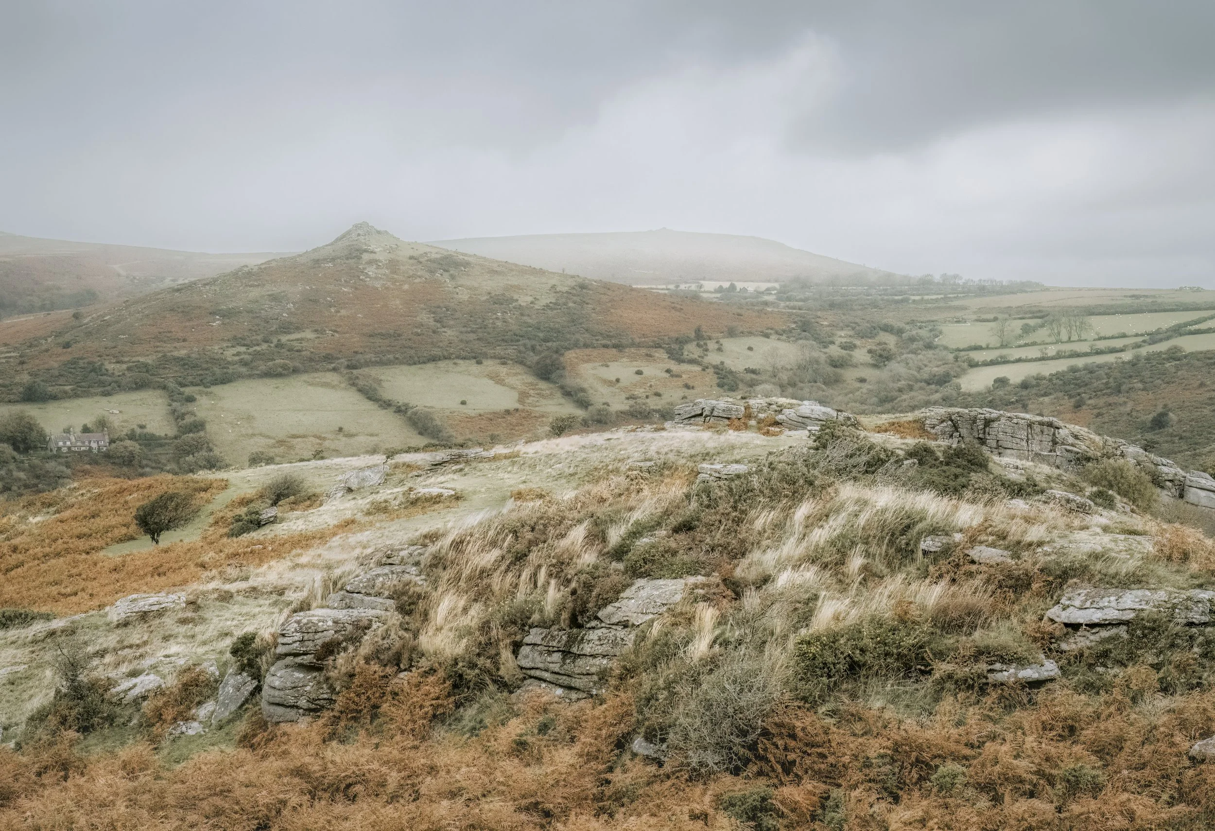 A scenic landscape of rolling hills with patches of grass and shrubs under a cloudy sky, featuring rocky outcrops and distant fields.