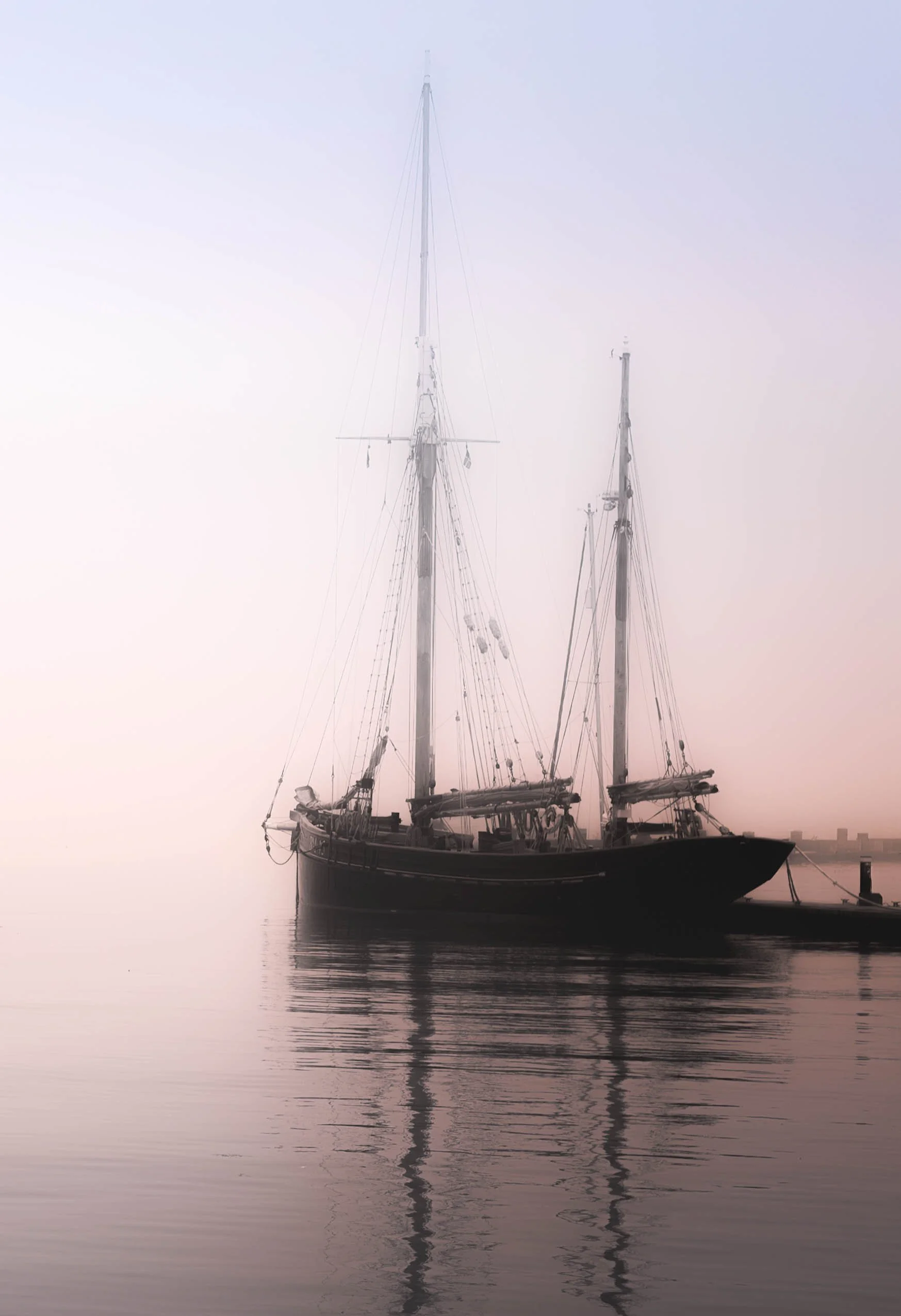 A black sailboat docked at a pier on calm water during sunset or sunrise, with soft pastel sky colors.