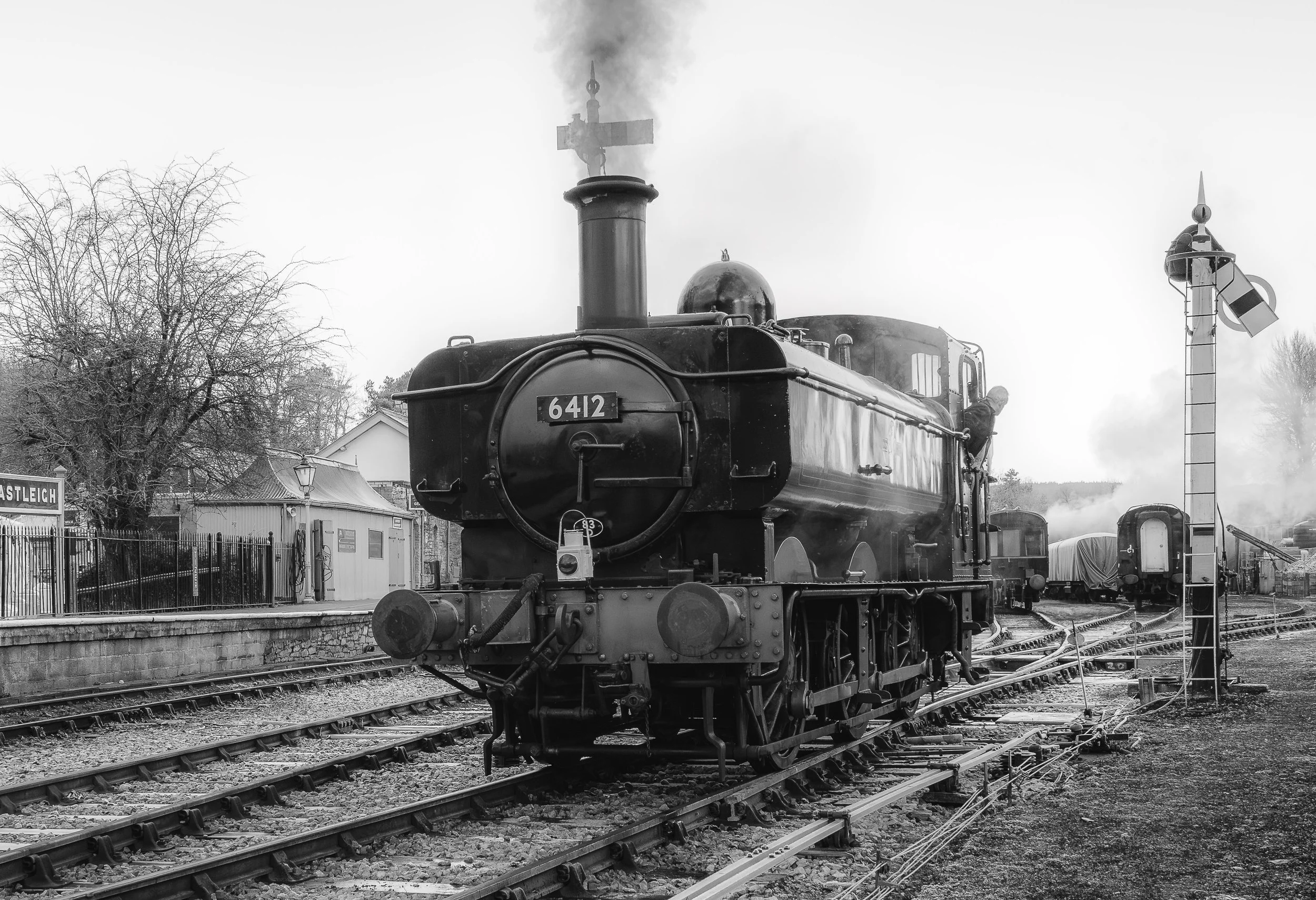 Black and white photo of a vintage steam train on railroad tracks with a signal light, trees, and buildings in the background.