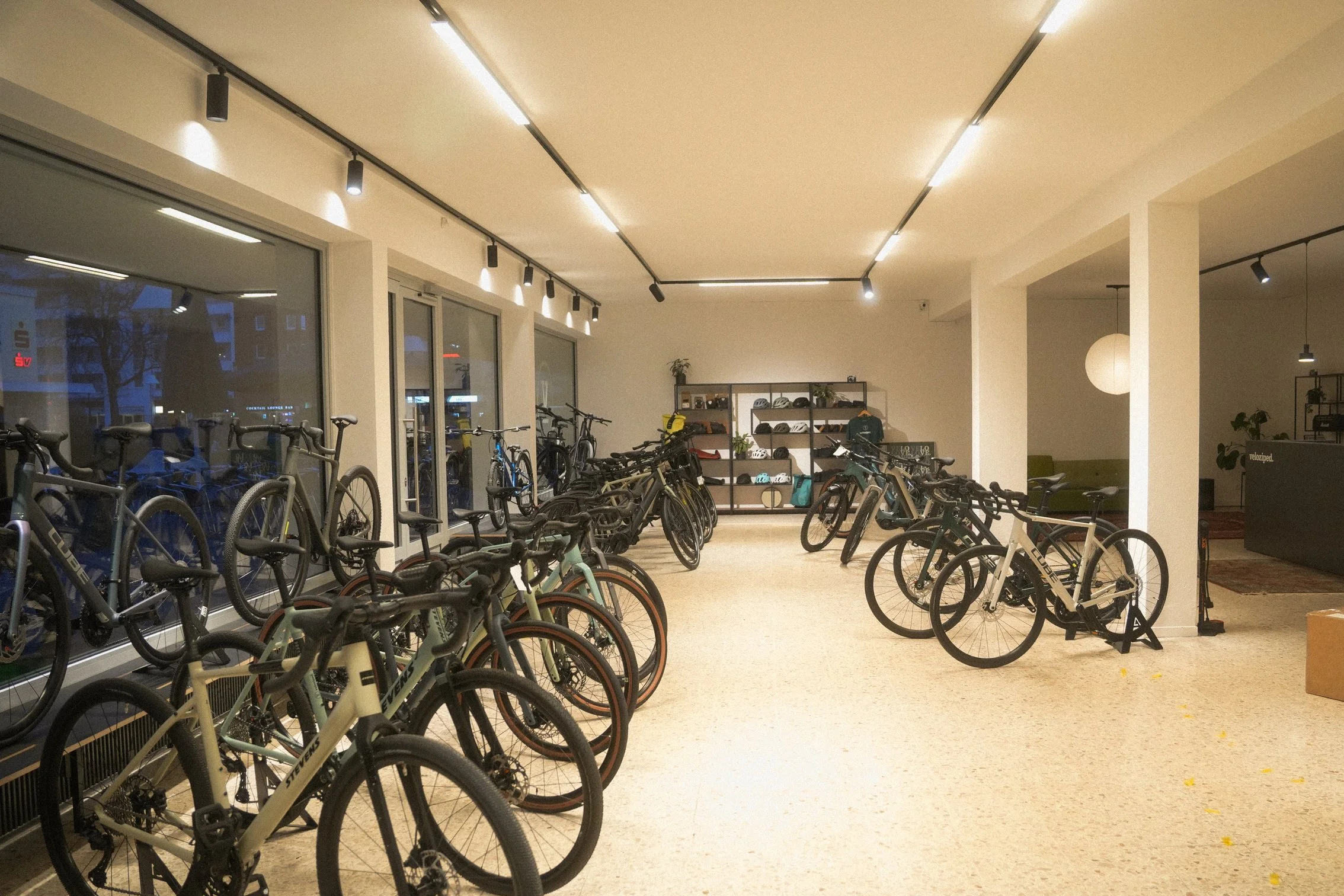 Innenaufnahme eines Fahrradgeschäfts oder einer Fahrradgarage mit mehreren abgestellten Fahrrädern, großen Fenstern und moderner Beleuchtung, sowie einer Regalwand im Hintergrund.