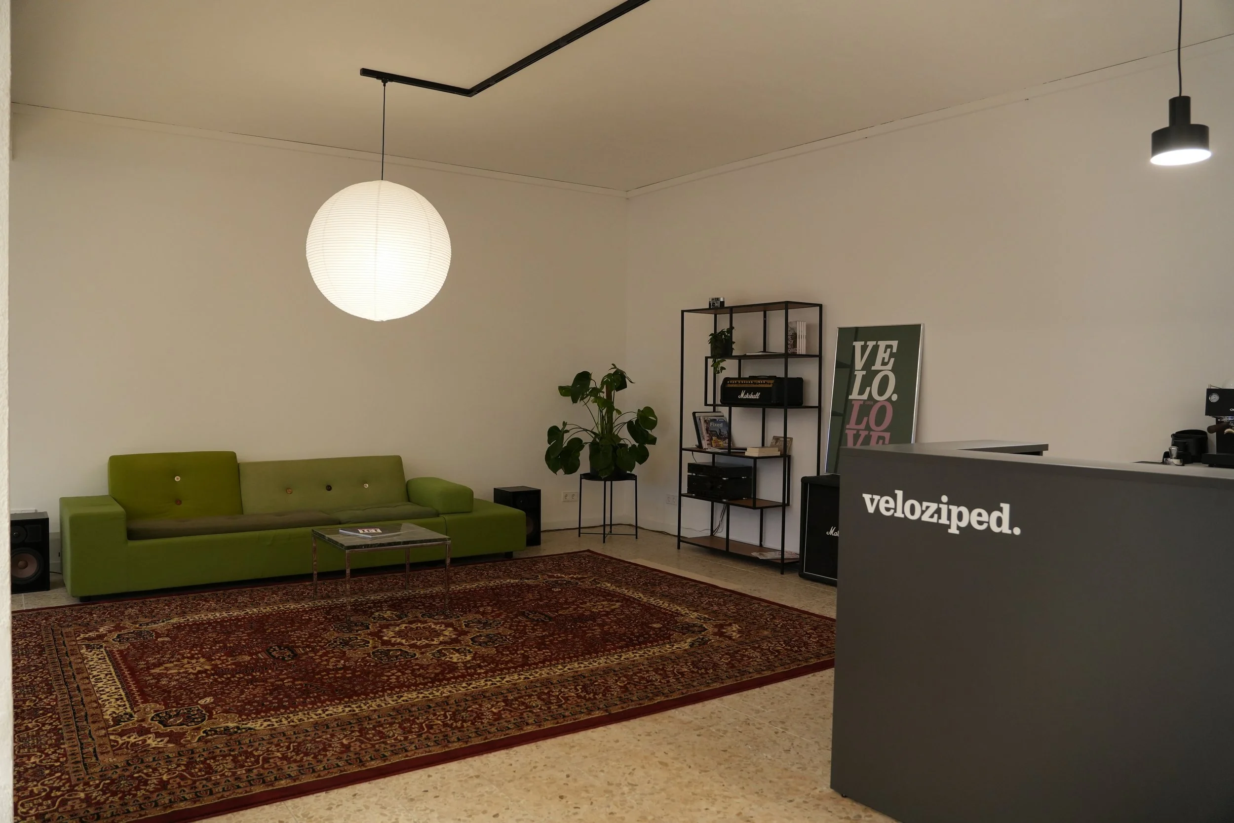 Ein modern eingerichteter Raum mit einem grünen Sofa, einem Regal mit Büchern und Dekorationsgegenständen, einer großen Wandkarte mit dem Wort „LOVE“ und einem Empfangstresen mit der Aufschrift „veloziped“.