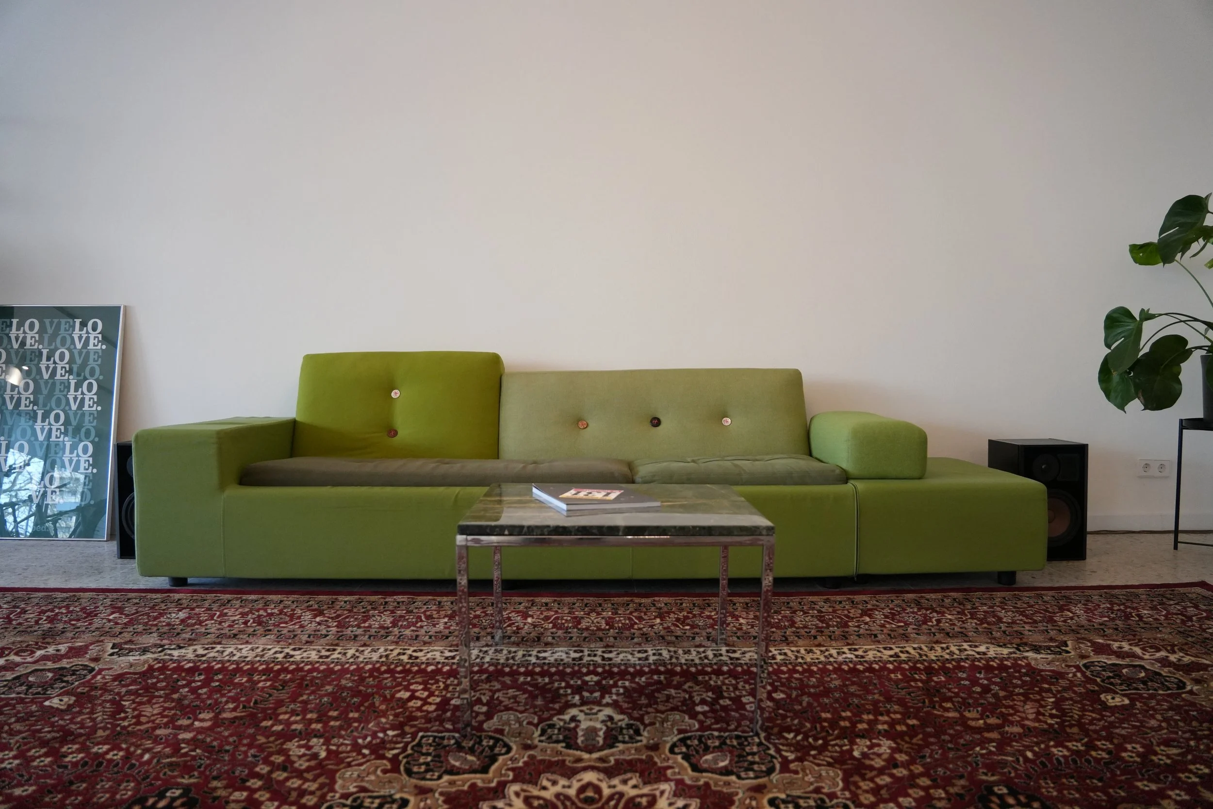 Ein modernes Wohnzimmer mit einem grünen Sofa, einem Marmortisch, einem großen Teppich mit Rot-, Schwarz- und Cremetönen, und einigen Dekorationsstücken, darunter ein gerahmtes Bild und eine Pflanze.