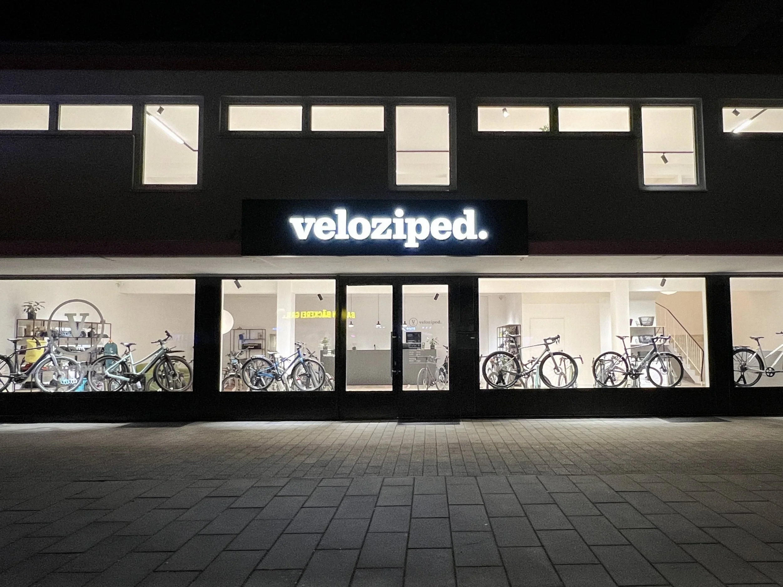 Anzeige eines Fahrradgeschäfts namens veloziped. mit Fahrrädern im Schaufenster bei Nacht.