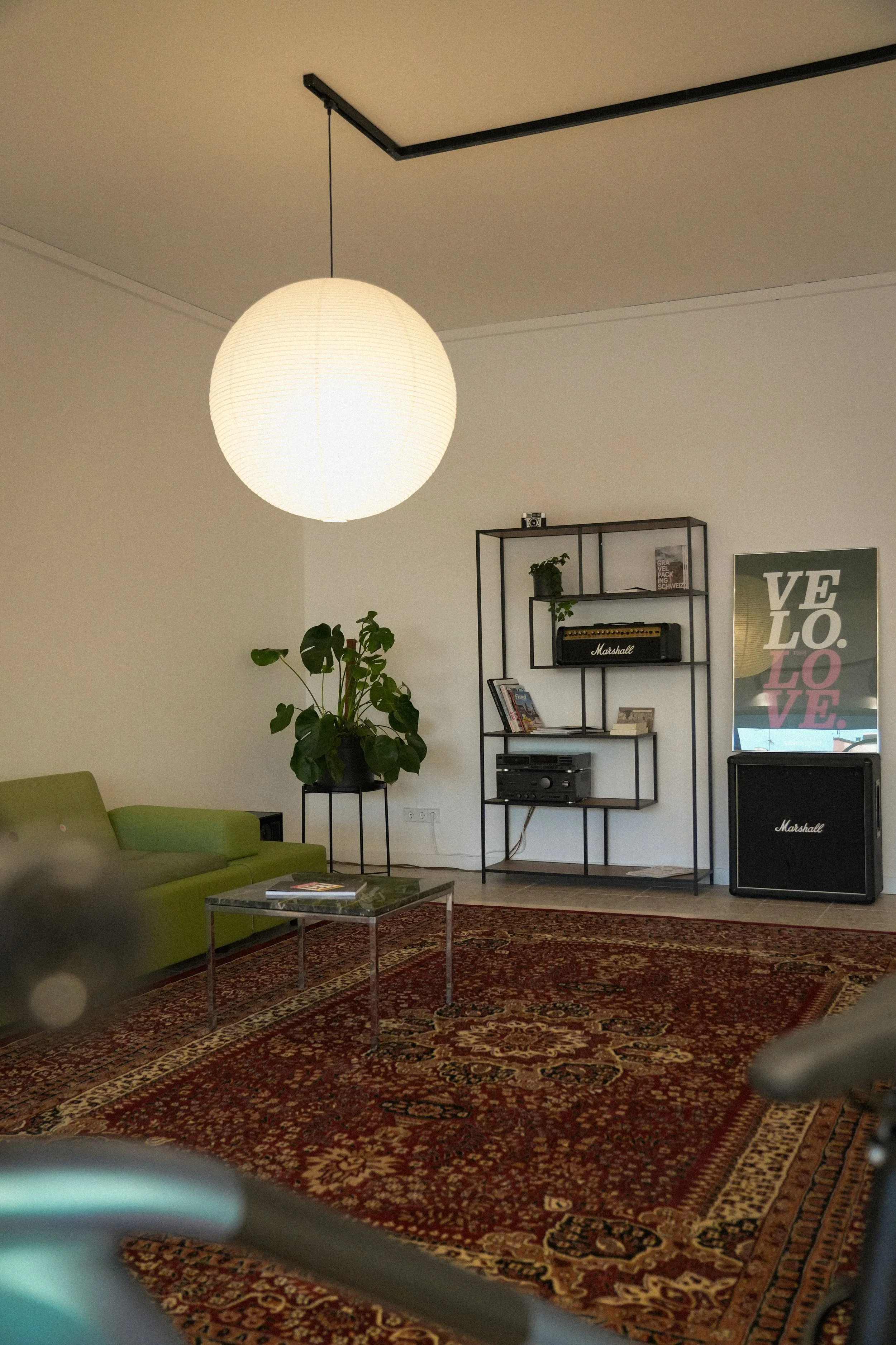 Ein Wohnzimmer mit grüner Couch, einem schwarzen Regal, einer großen Topfpflanze, einem großen weißen Papierlampenschirm, einem roten Perserteppich, einem schwarzen Lautsprecher und einem Poster mit den Worten »VELO LOVE«.