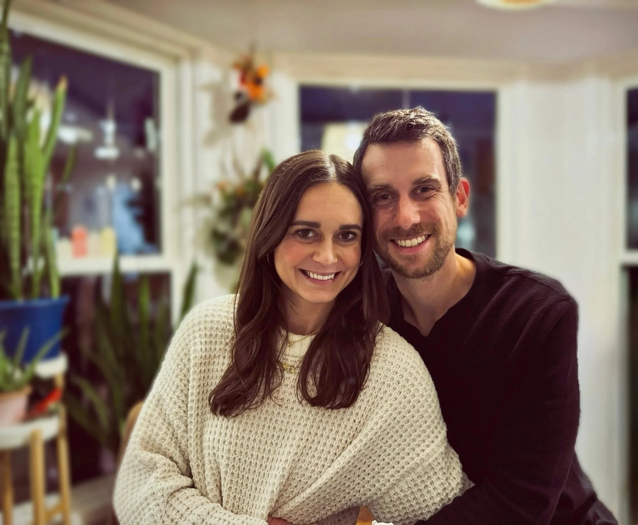A smiling woman and man sitting close together in a cozy indoor setting, with large windows and houseplants in the background.