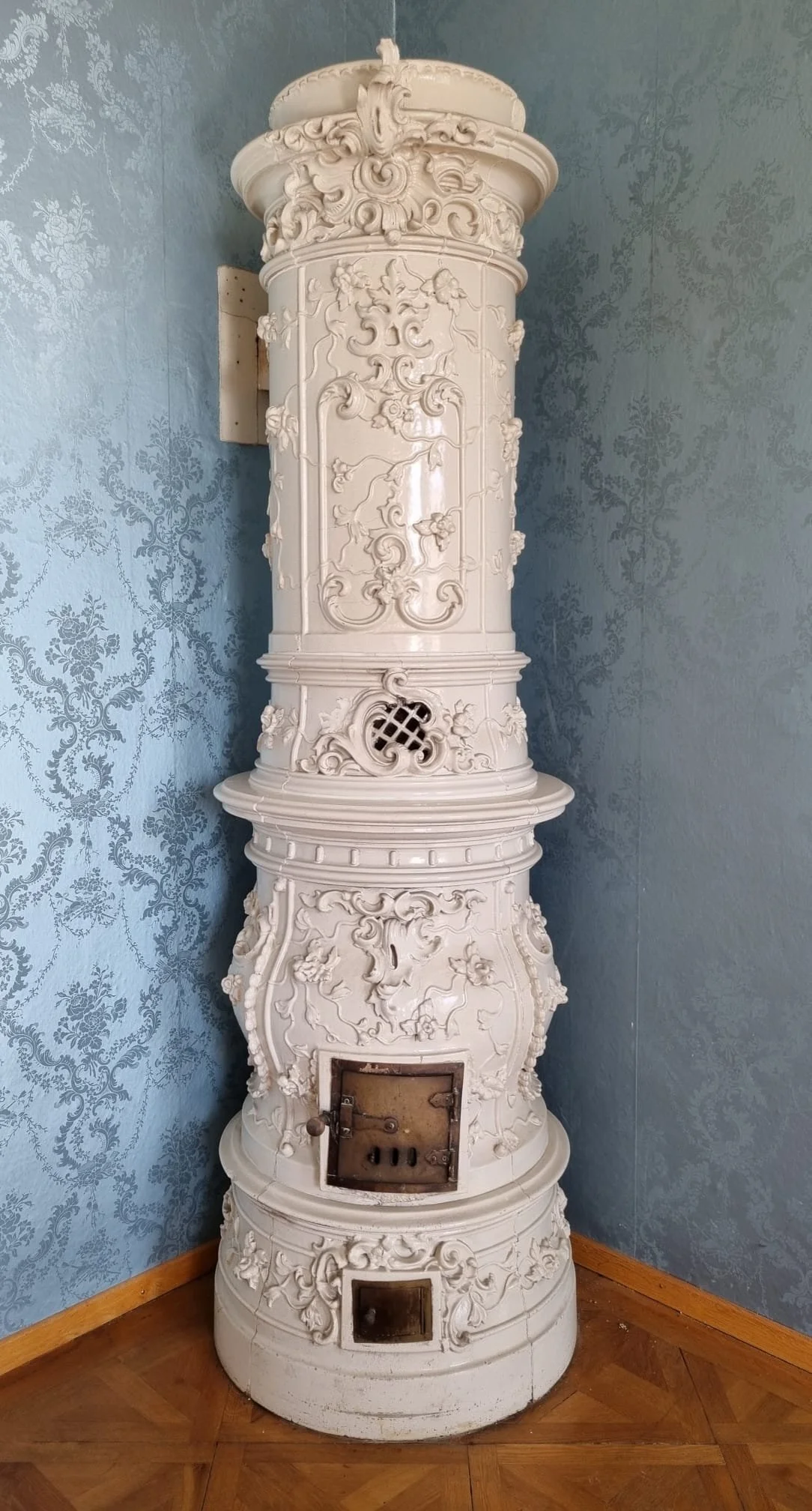 Ein weißer, kunstvoll verzierter Kamin mit floralen und barockartigen Mustern in einem Raum mit blauer Tapete und Holzparkett.