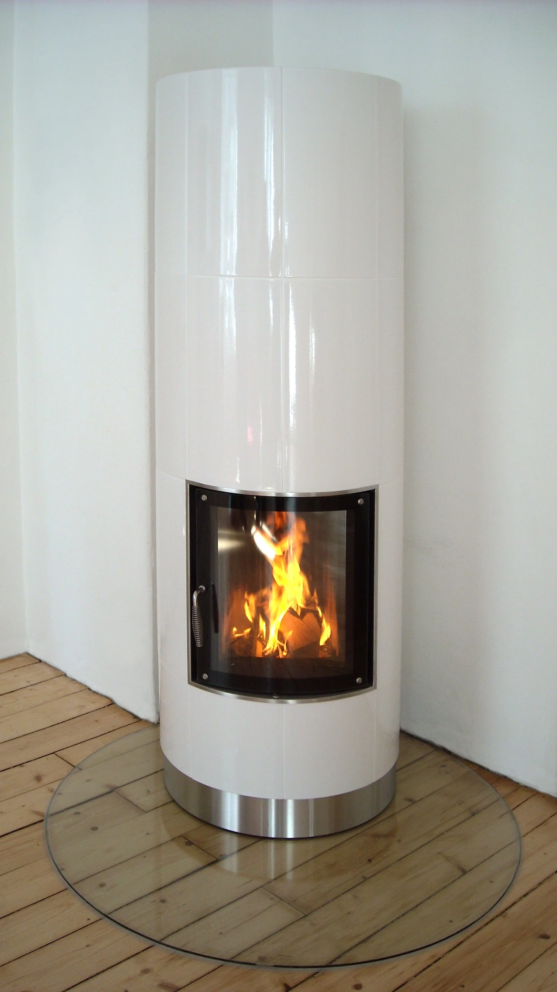 Ein moderner, weißer Kamin mit Glasfront, in dem ein Feuer brennt, auf Parkettboden vor weißer Wand.