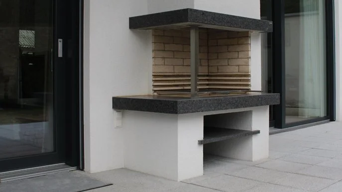 Ein moderner Außenkamin mit schwarzer Granitplatte, einer Mauer aus Ziegelsteinen und einer weißen Sockelbasis, neben einer Glasschiebetür.