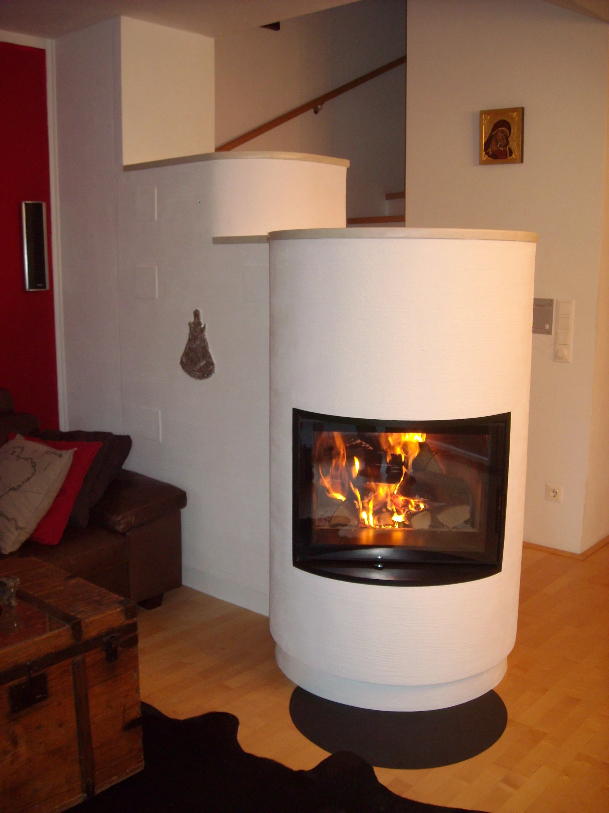 Ein moderner, runder Kamin mit Feuer in einem Wohnzimmer, neben einem Sofa mit Kissen, auf dunklem Teppich vor hellem Holzboden, mit Kunst an der Wand.