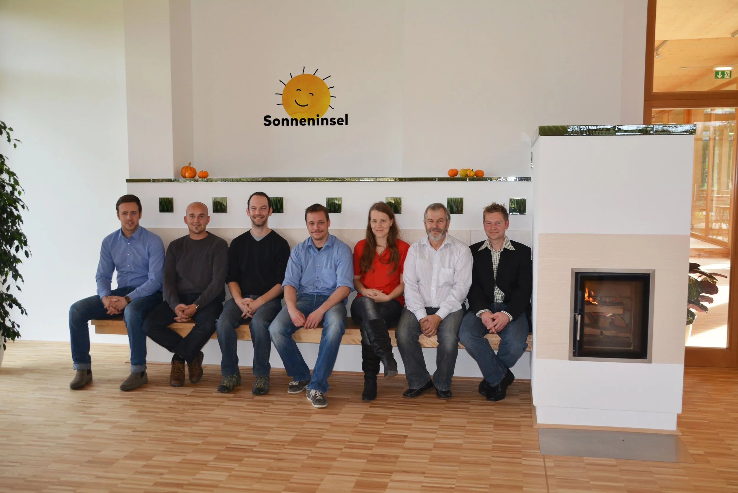 Sieben Personen sitzen auf einer Bank in einem modernen Raum mit Kamin, hinter ihnen an der Wand ein Sonnen-Logo und der Schriftzug 'Sonneninsel'.