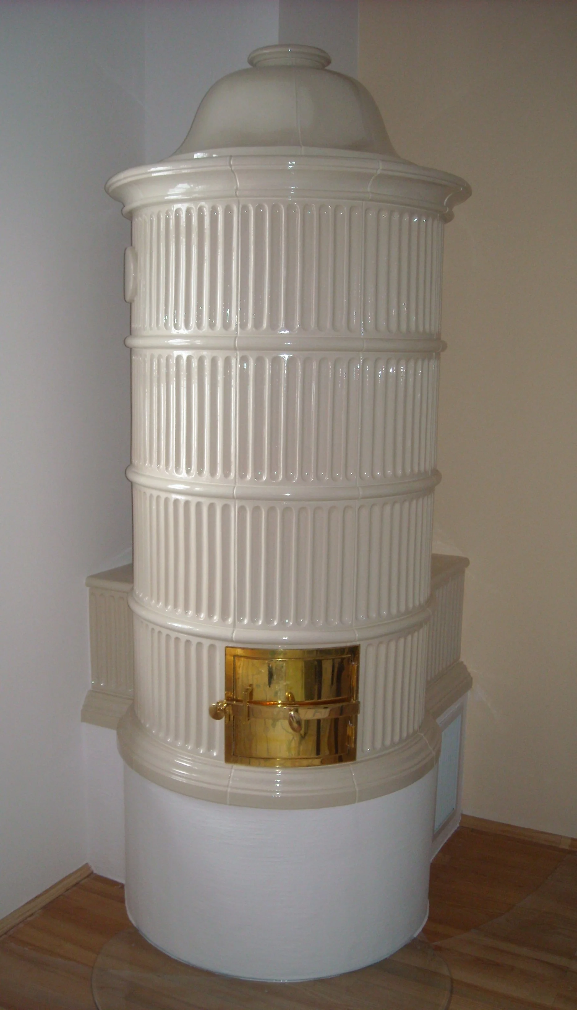 Ein weißer, runder Kachelofen mit goldener Klappe, auf Holzfußboden in einem Raum.