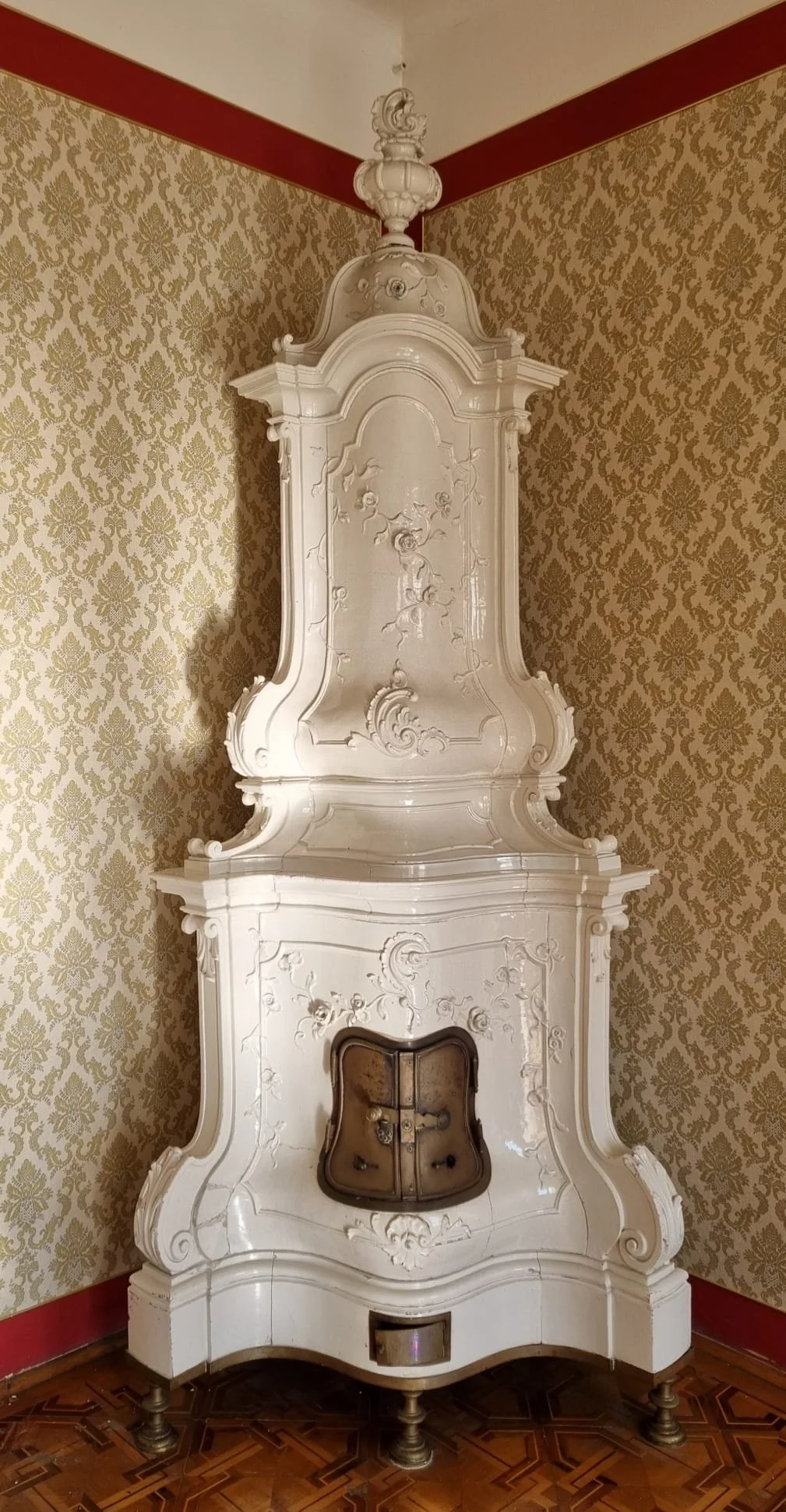 Antiker weißer Ofen mit kunstvollen Verzierungen in einem Raum mit gold- und beige-gemusterten Tapete und Holzboden.