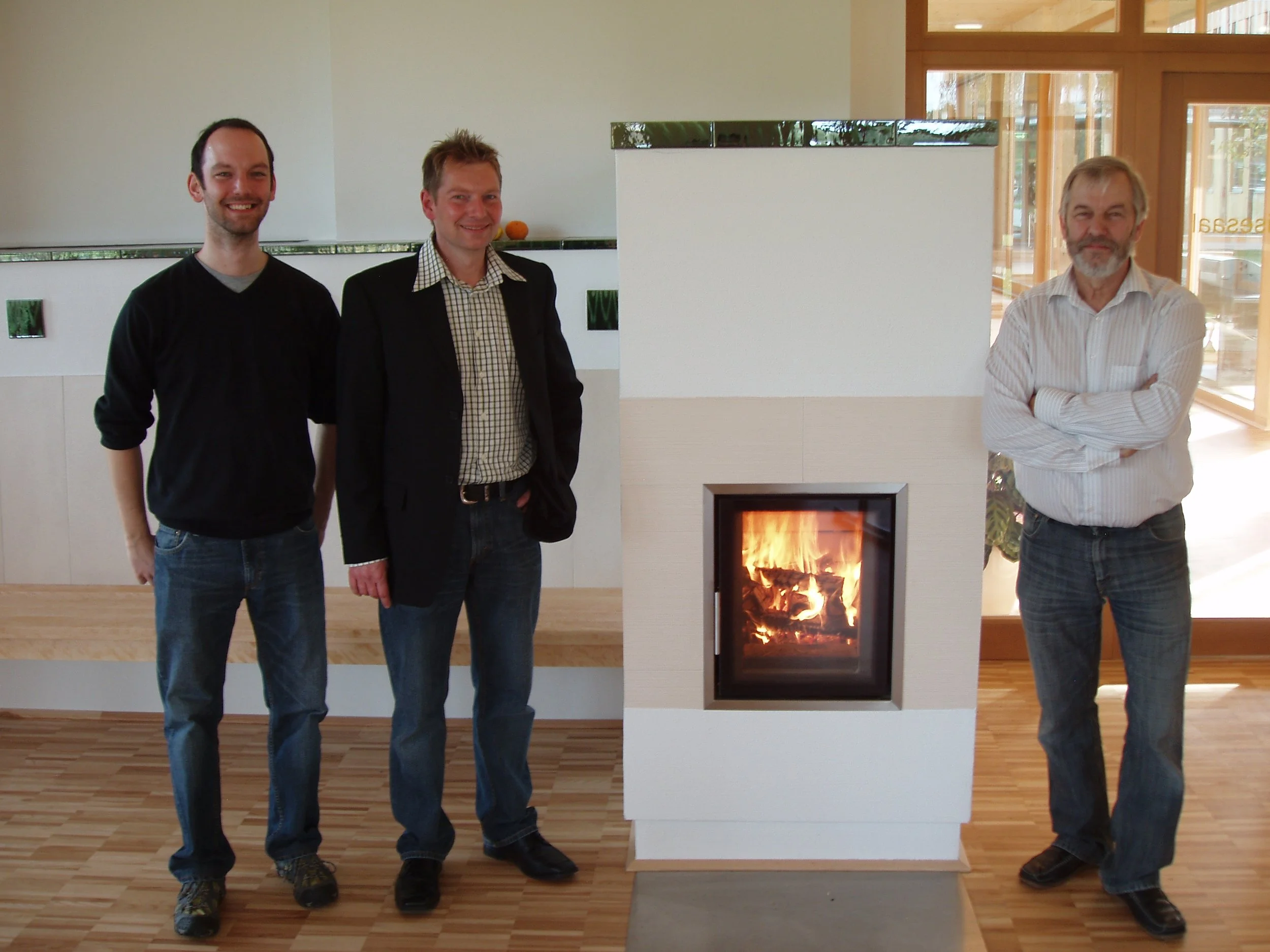 Drei Männer stehen neben einem modernen Kamin mit Feuer, in einem Raum mit Holzfußboden und großen Fenstern, die Tageslicht hineinlassen.