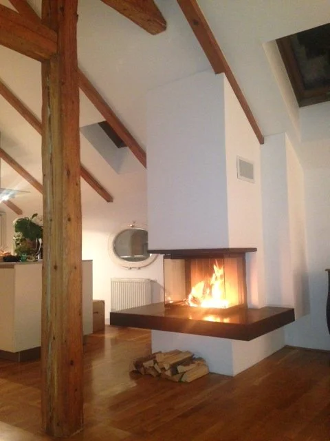 Moderner Kamin im Wohnzimmer mit großem Sichtfenster, sichtbare Holzbalken an der Decke.
