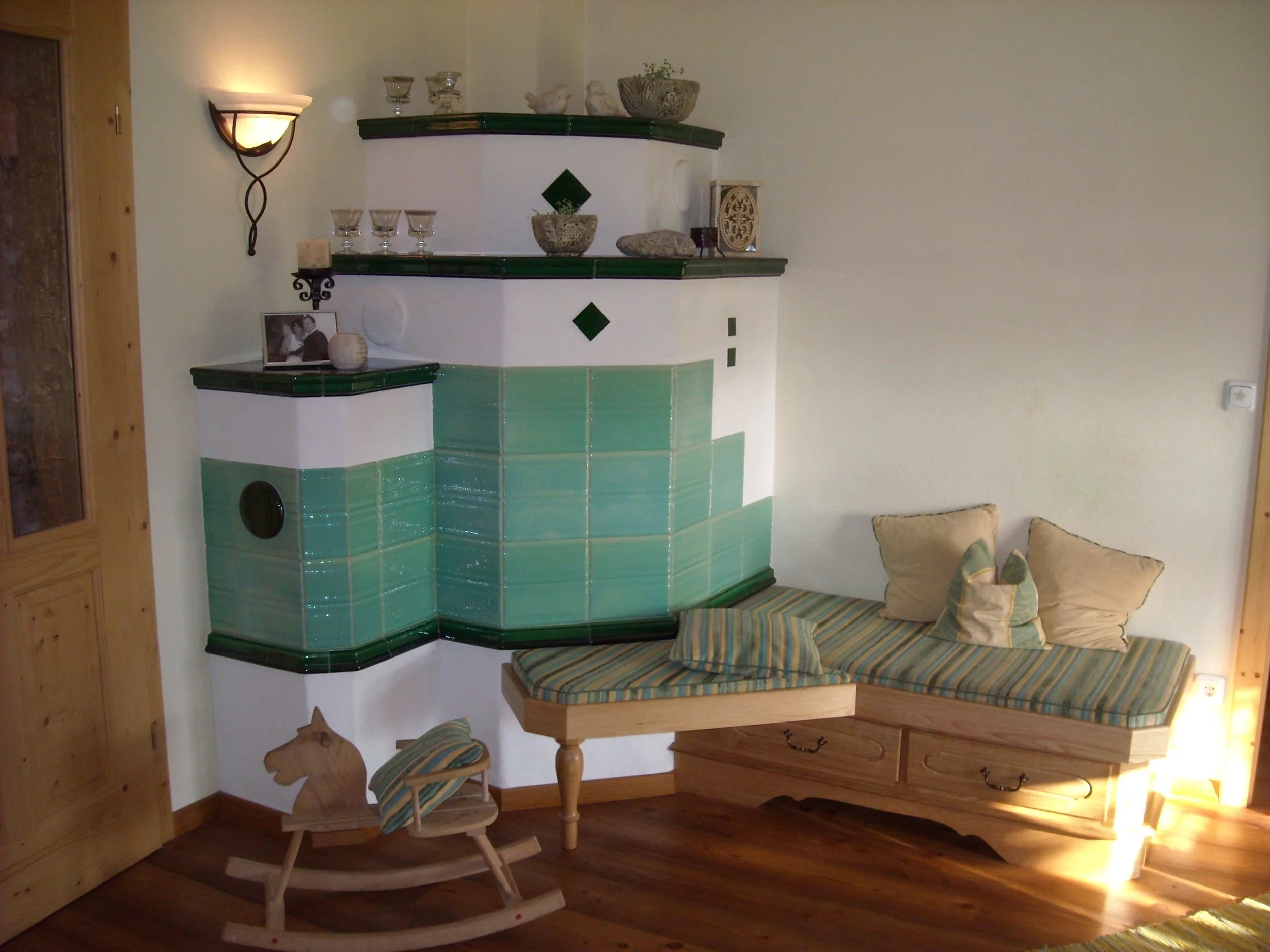 Rustikaler Kaminofen mit grün-glasierten Kacheln, gemütlicher Holzbank mit Kissen, Holzpferd-Schaukel und dekorativen Gegenständen.