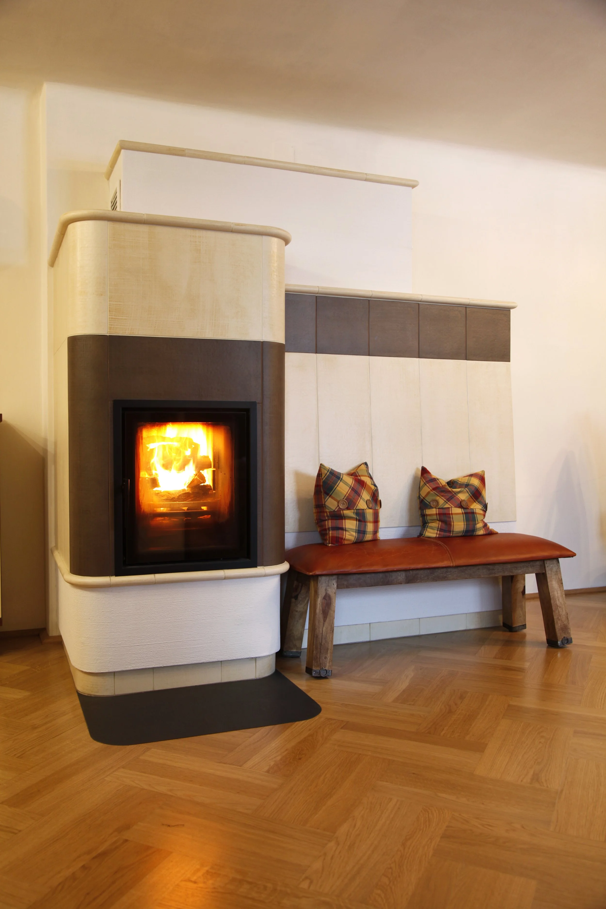 Ein stilvoller Kamin in weiß-grau gehalten, mit Feuer, daneben eine Holzbank mit zwei gepolsterten Kissen, auf einem Holzboden in einem modernen Raum.