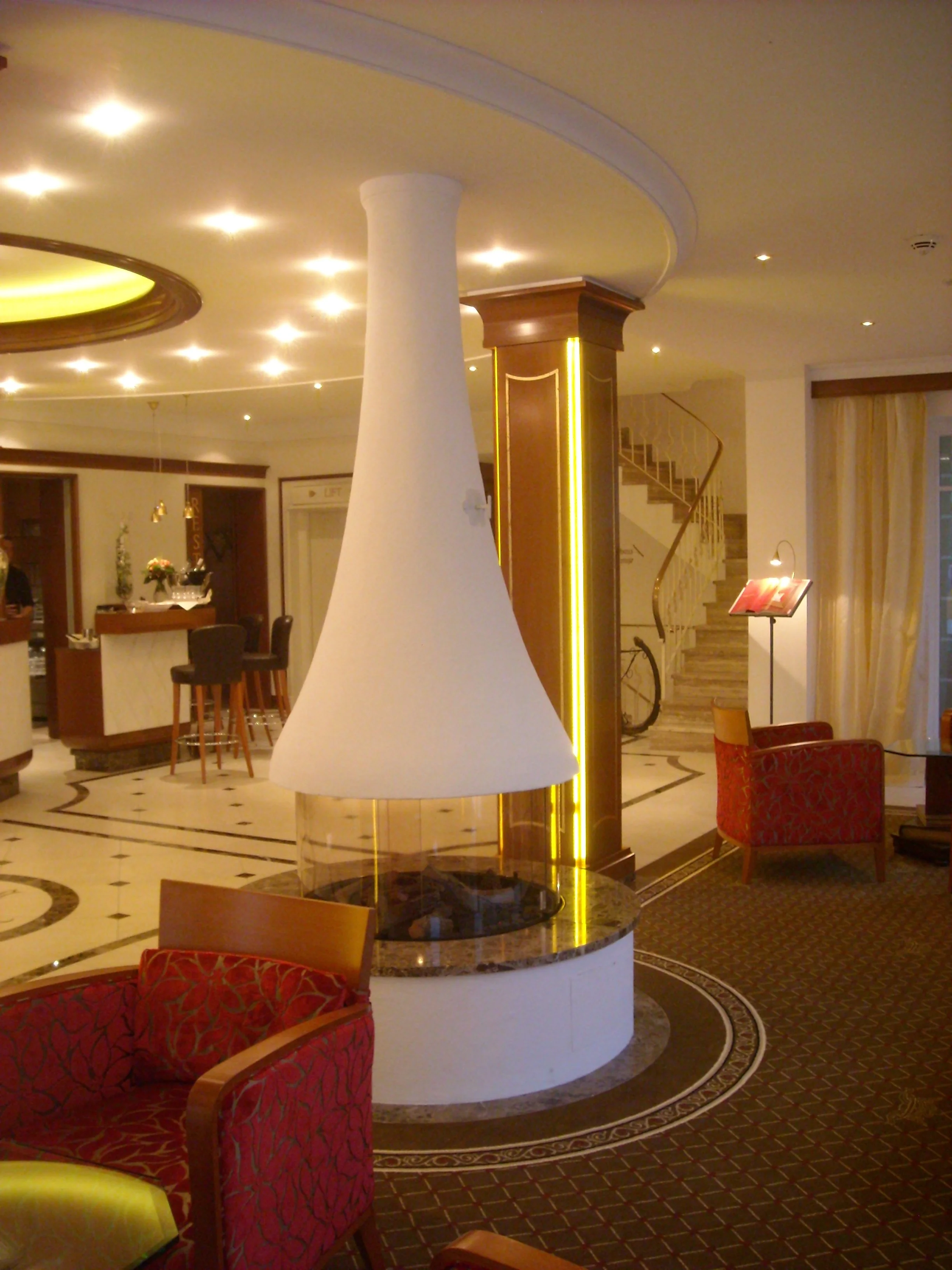 Innenaufnahme einer Hotellobby mit einladendem Kamin, roten Sesseln, Treppe und Empfangstresen.