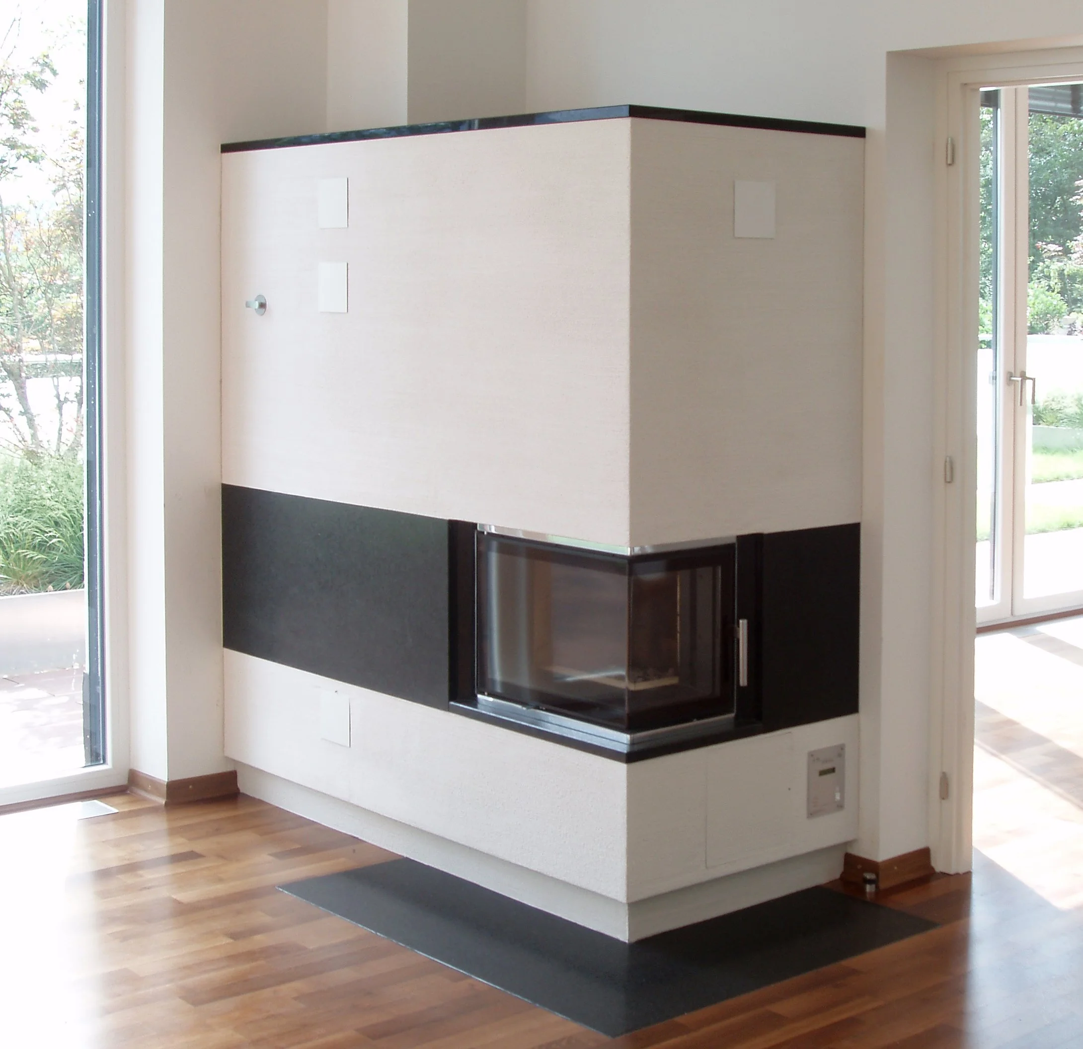 Moderner eingebaute Kamin in einem hellen Wohnzimmer, mit Holzfußboden und Glasfenster im Hintergrund.