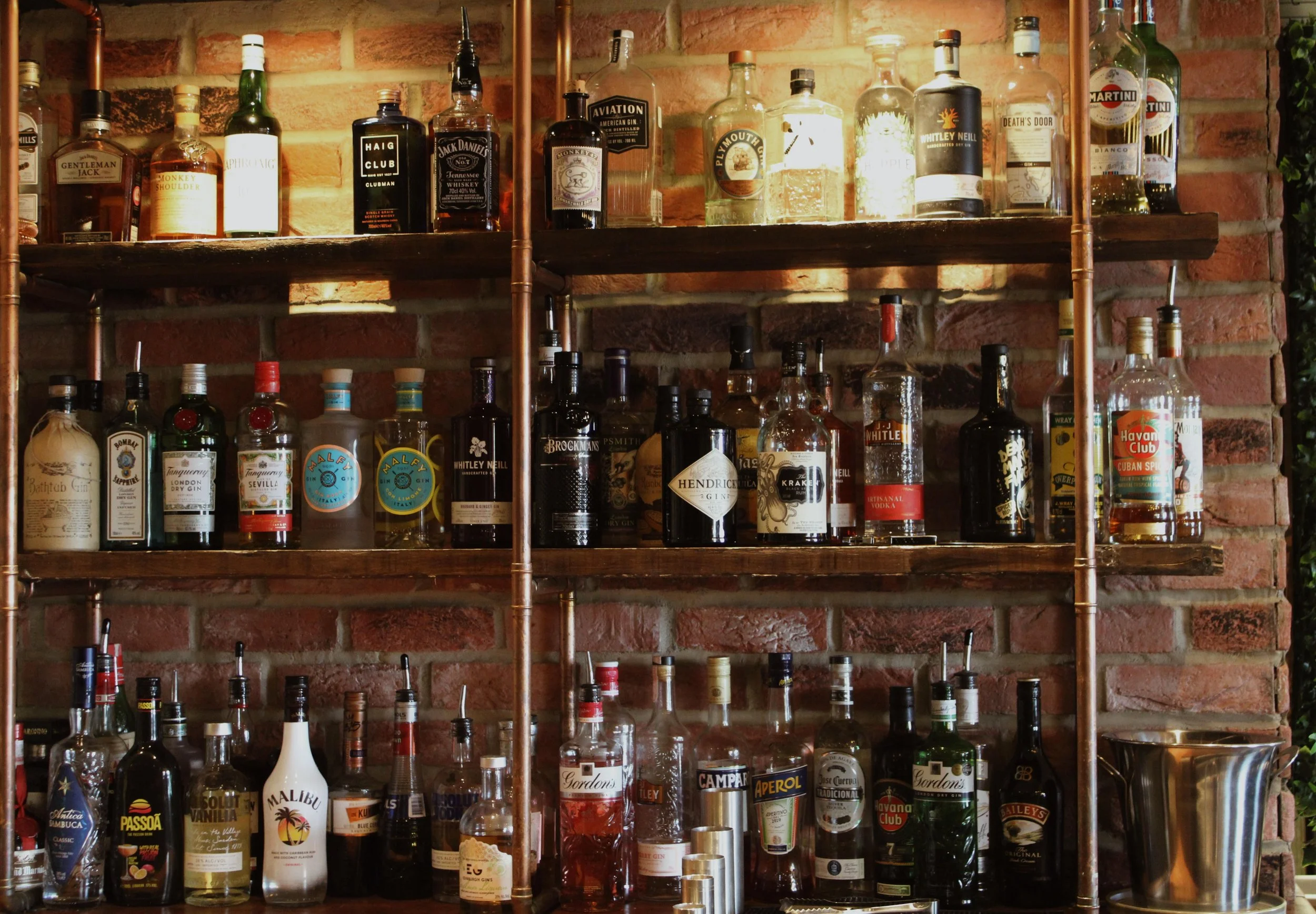 A wooden bar shelf mounted against a brick wall, holding various bottles of liquor including whiskey, gin, vodka, and rum, with some bottles featuring recognizable labels such as Malibu and Havana Club.