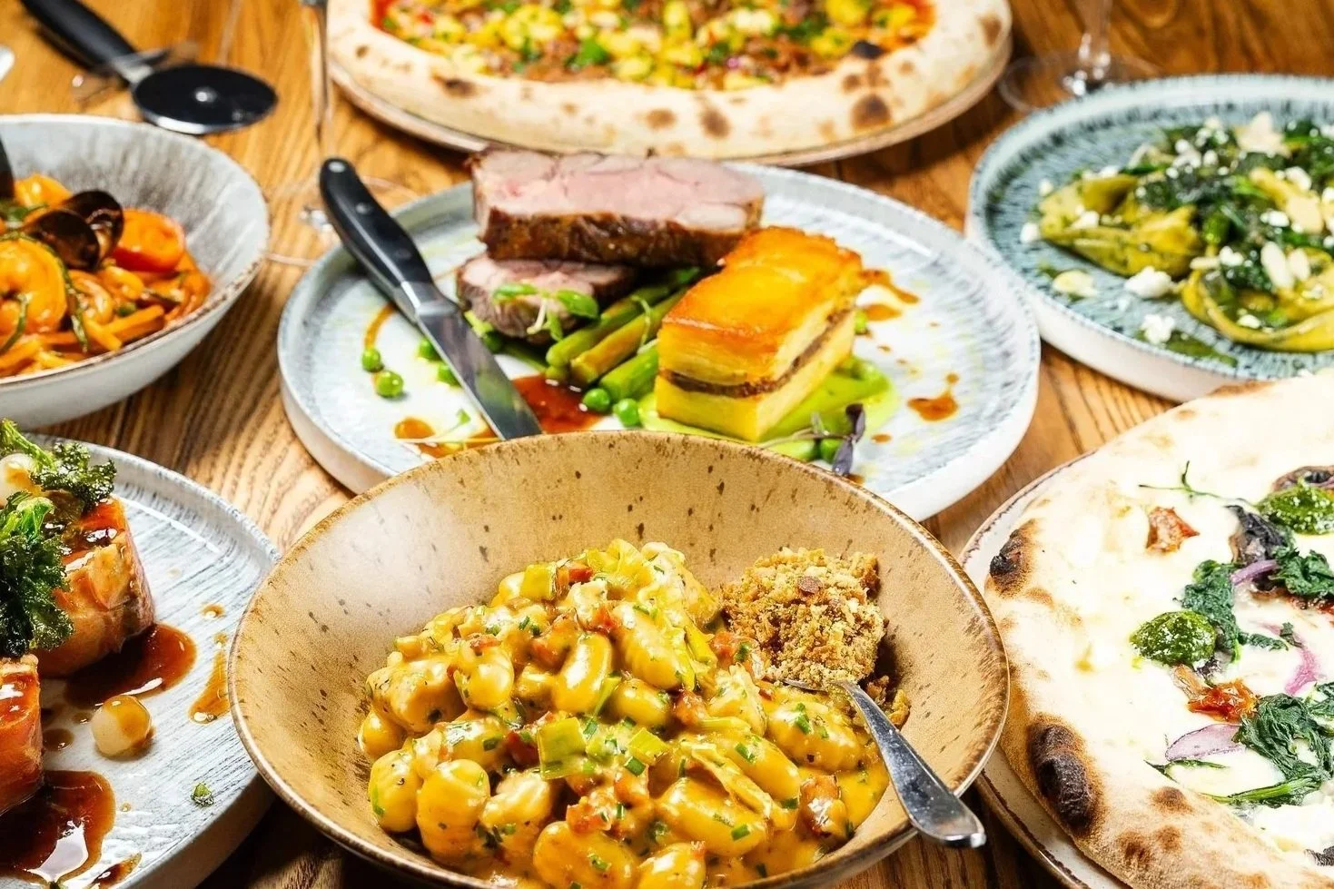 Various Italian dishes on a wooden table, including pizza, pasta, and meat with vegetables.