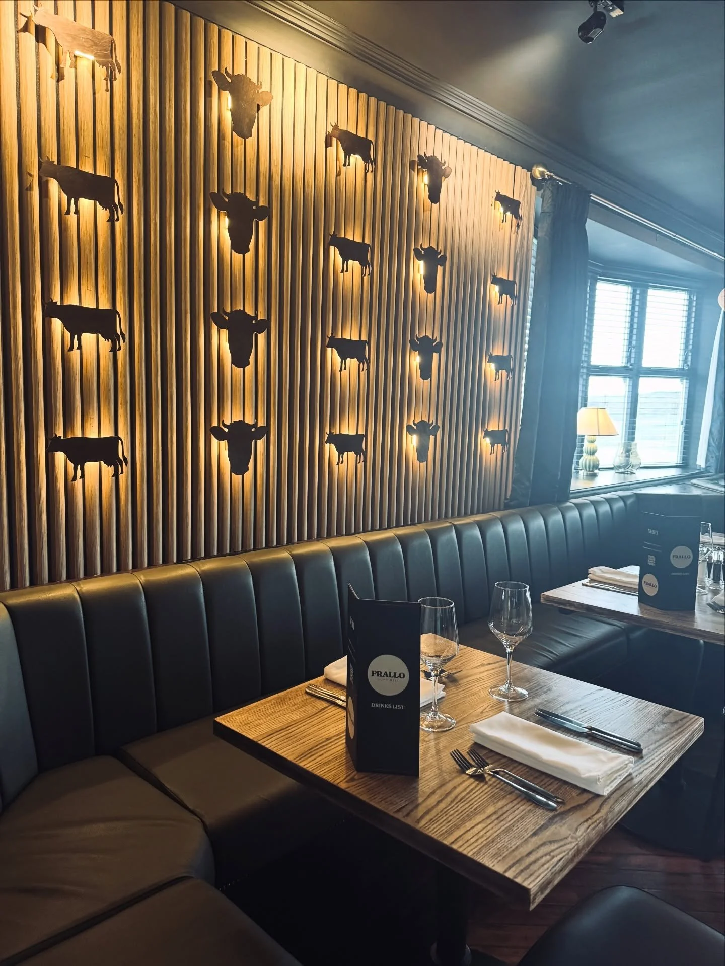Restaurant interior with a wooden wall decorated with illuminated cow and bull head silhouettes, dark leather seating, wooden tables set with napkins, glassware, and menus, near a window with blinds and a lamp.