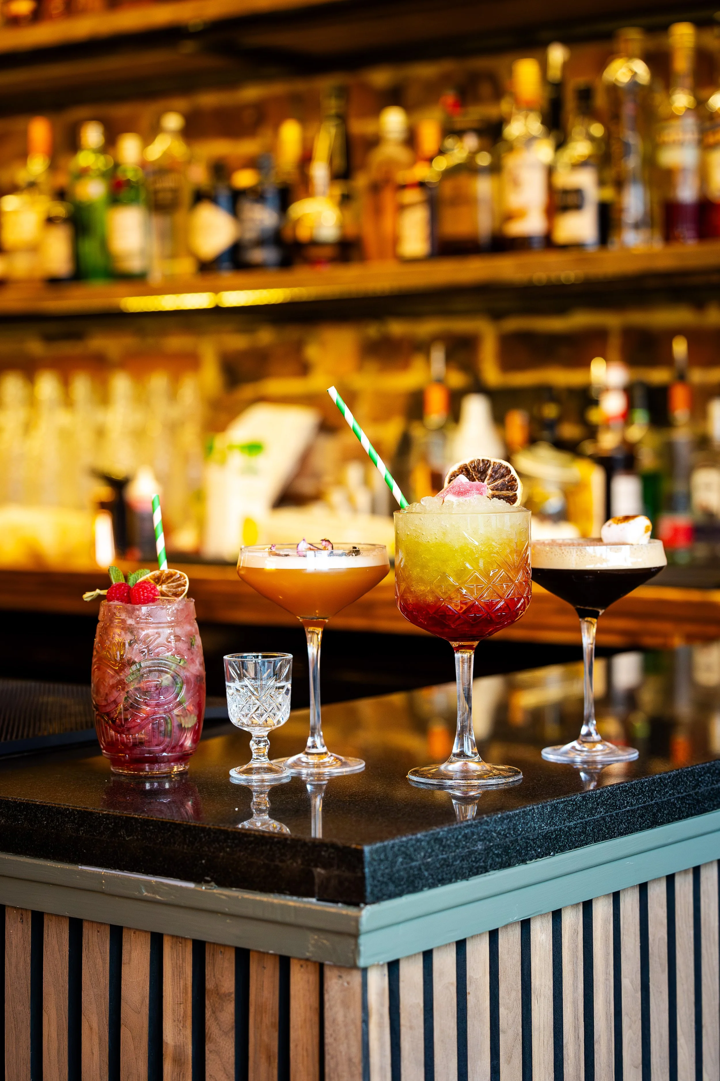 Four colorful cocktails on a bar counter with a blurred bar back in the background.