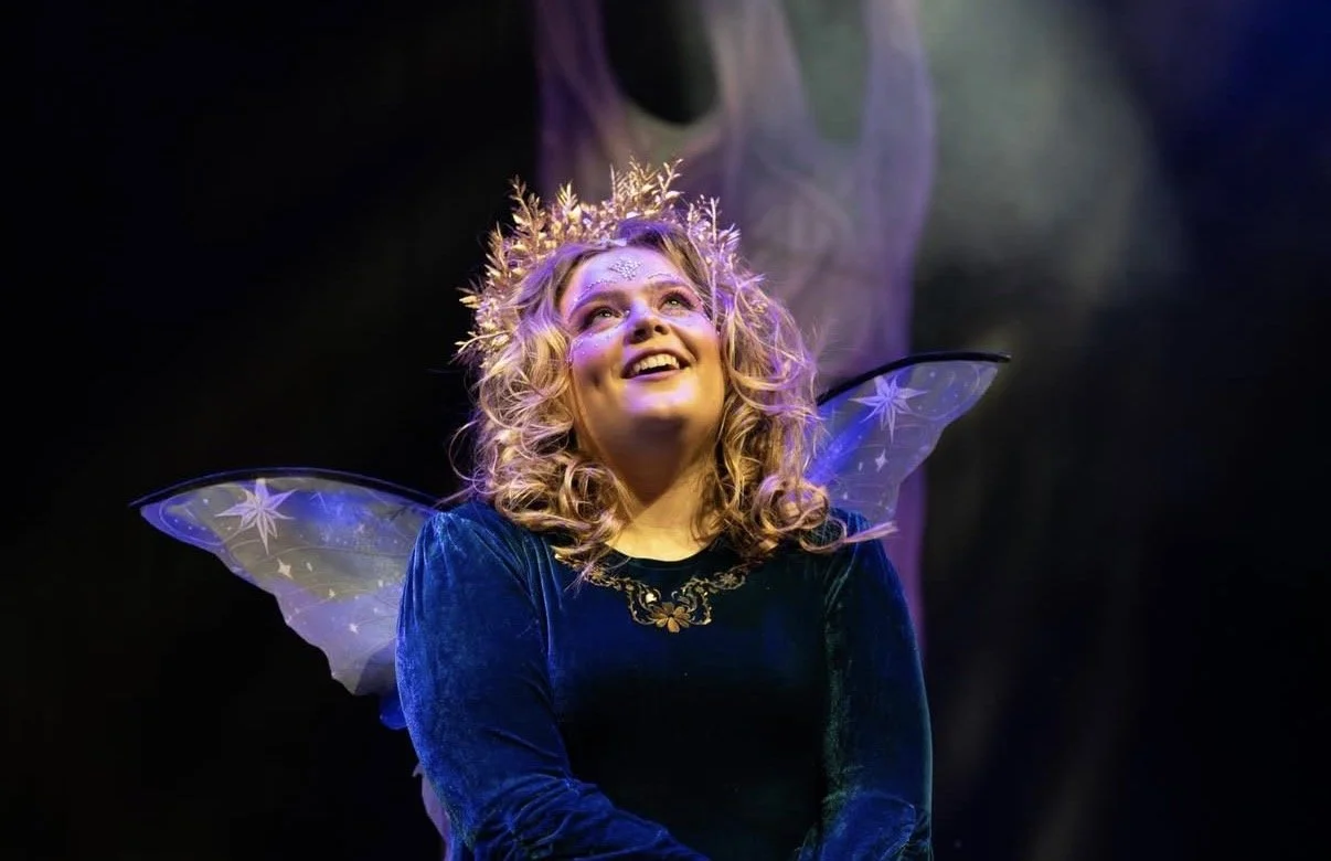 A woman with short hair wearing a leather jacket and black headband with a bow, illuminated by green and yellow stage lights, holding a cane or scepter, with a serious expression.