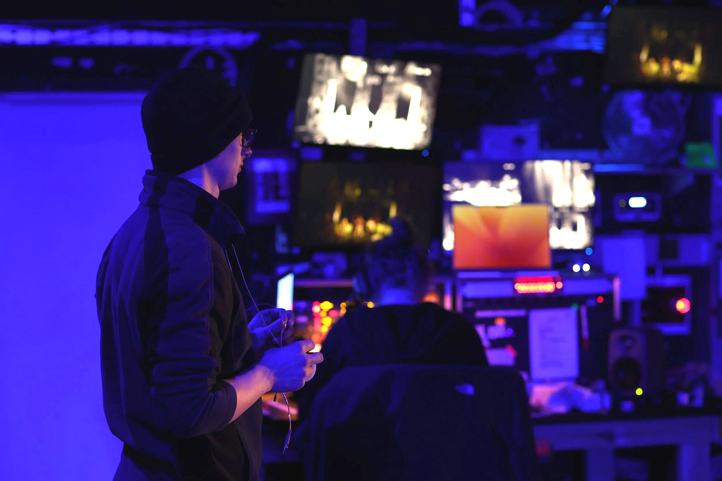 A person wearing a red winter hat with a pom-pom, glasses, and headphones is working at a DJ station. There is a large screen behind them displaying the text 'GREEN STREET Celebration' and a laptop in front with audio editing software.