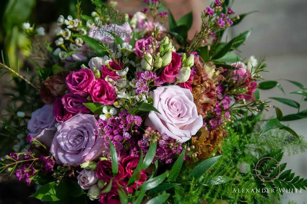 Bride in a strapless white wedding gown holding a bouquet of white roses and greenery, standing beside groom in a blue suit at an outdoor wedding.