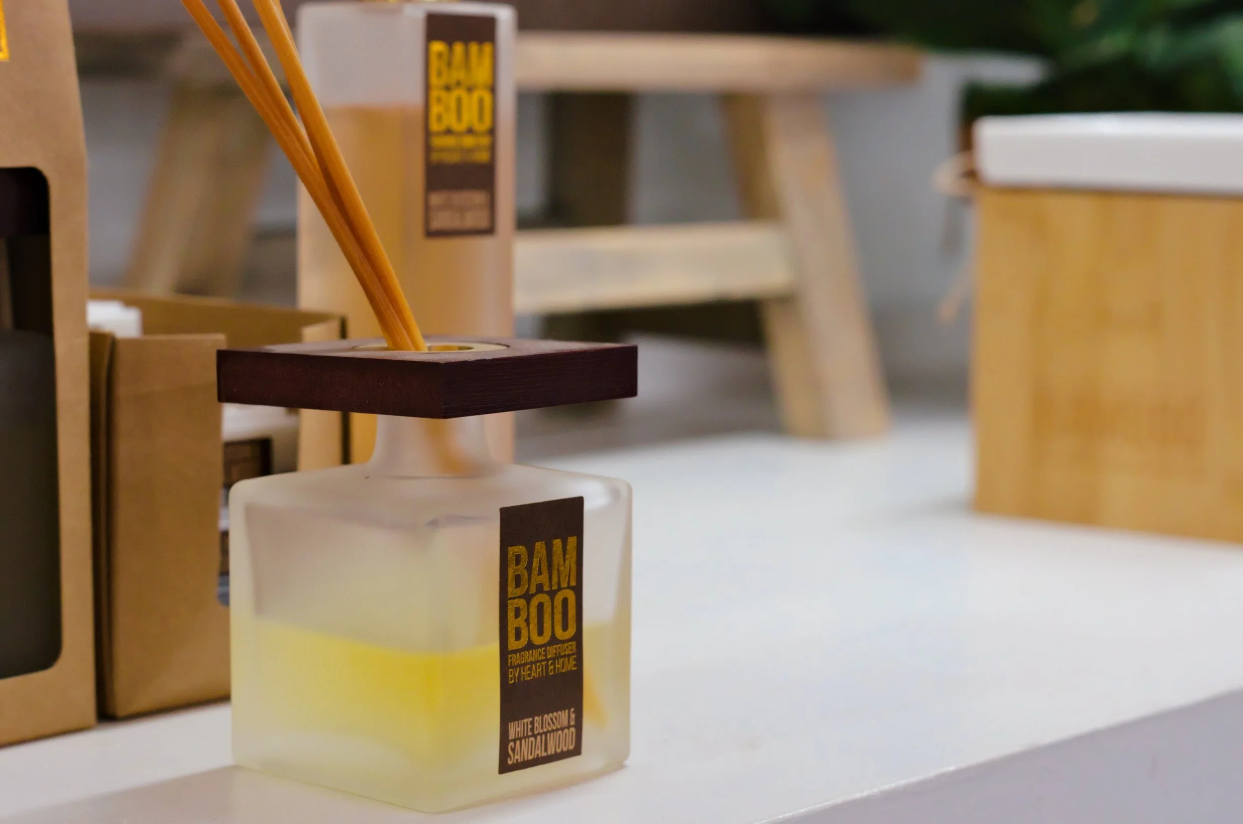A fragrance diffuser labeled BAMBOO with bamboo sticks in a glass container, placed on a white surface with wooden furniture and containers in the background.